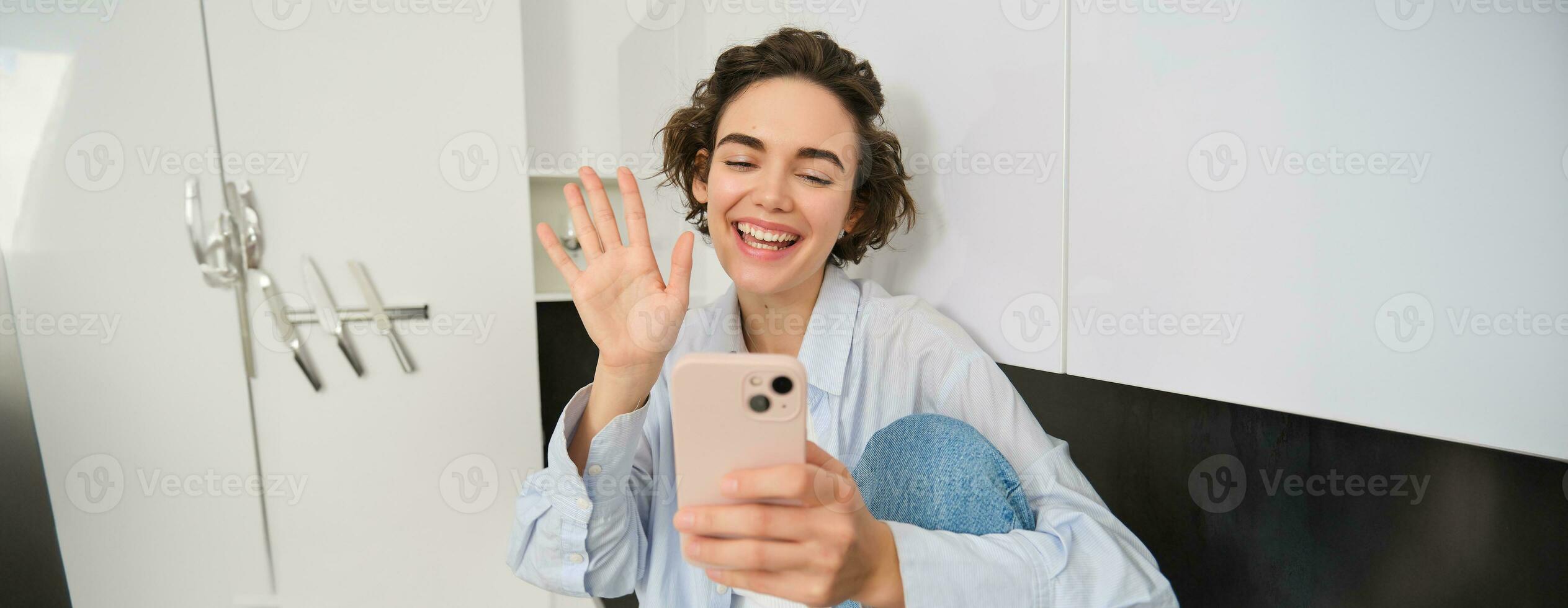 retrato de sonriente simpático mujer, mirando a móvil teléfono, olas mano a teléfono inteligente cámara, vídeo charlas, conecta a en línea conversación, se sienta a hogar y negociaciones con alguien foto