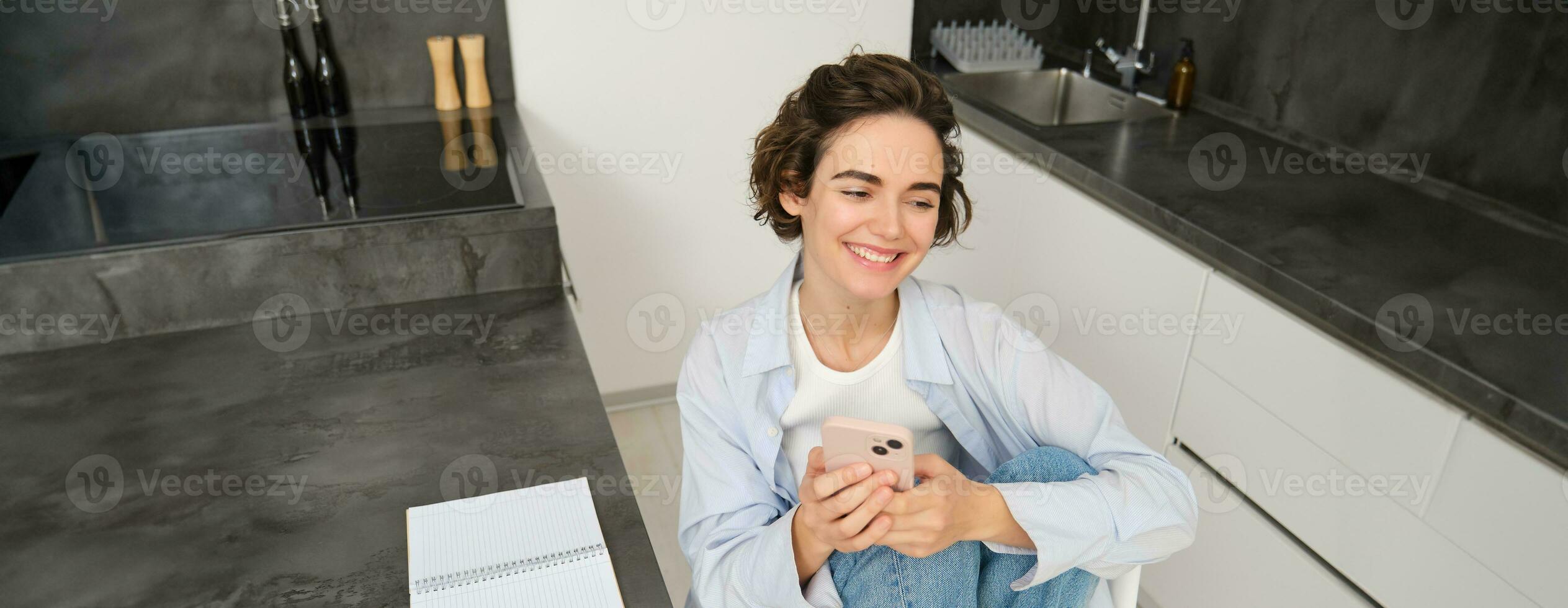 Young woman having break from work, sitting in kitchen and using smartphone, girl studies at home, sends a message on mobile phone photo
