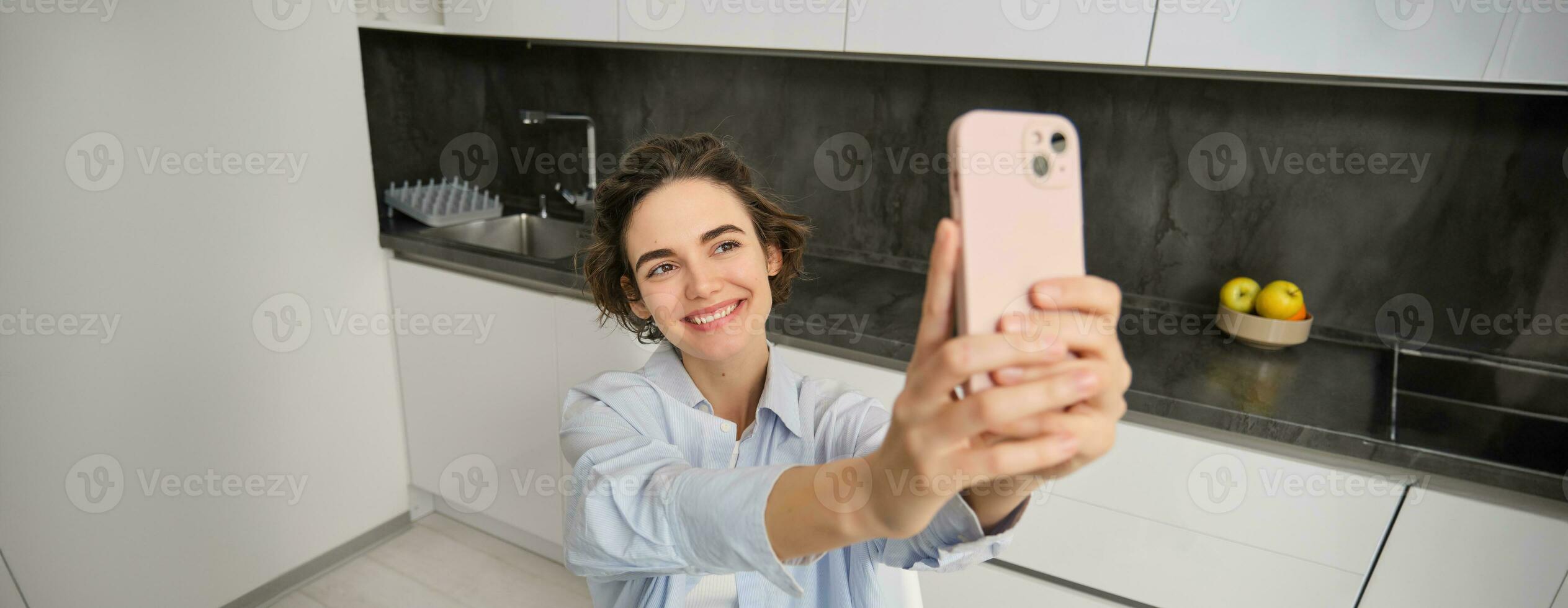 Stylish smiling girl takes selfie on smartphone at home, makes photo of herself in kitchen, poses for picture with mobile phone