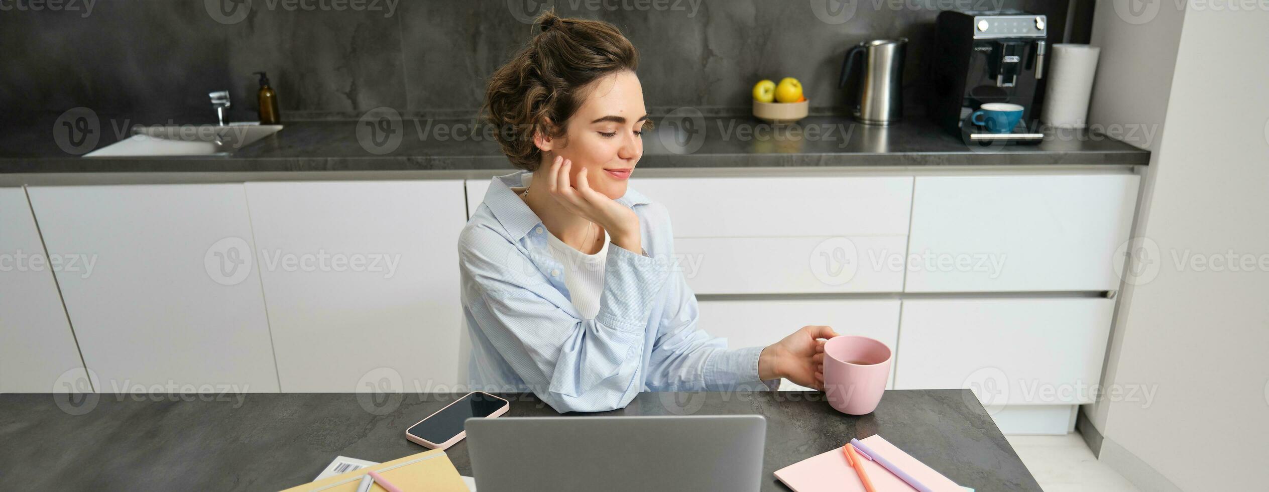 Beautiful woman managing her work from home, drinking coffee and looking at laptop, sitting in kitchen with paperwork, doing her taxes, studying indoors on remote photo
