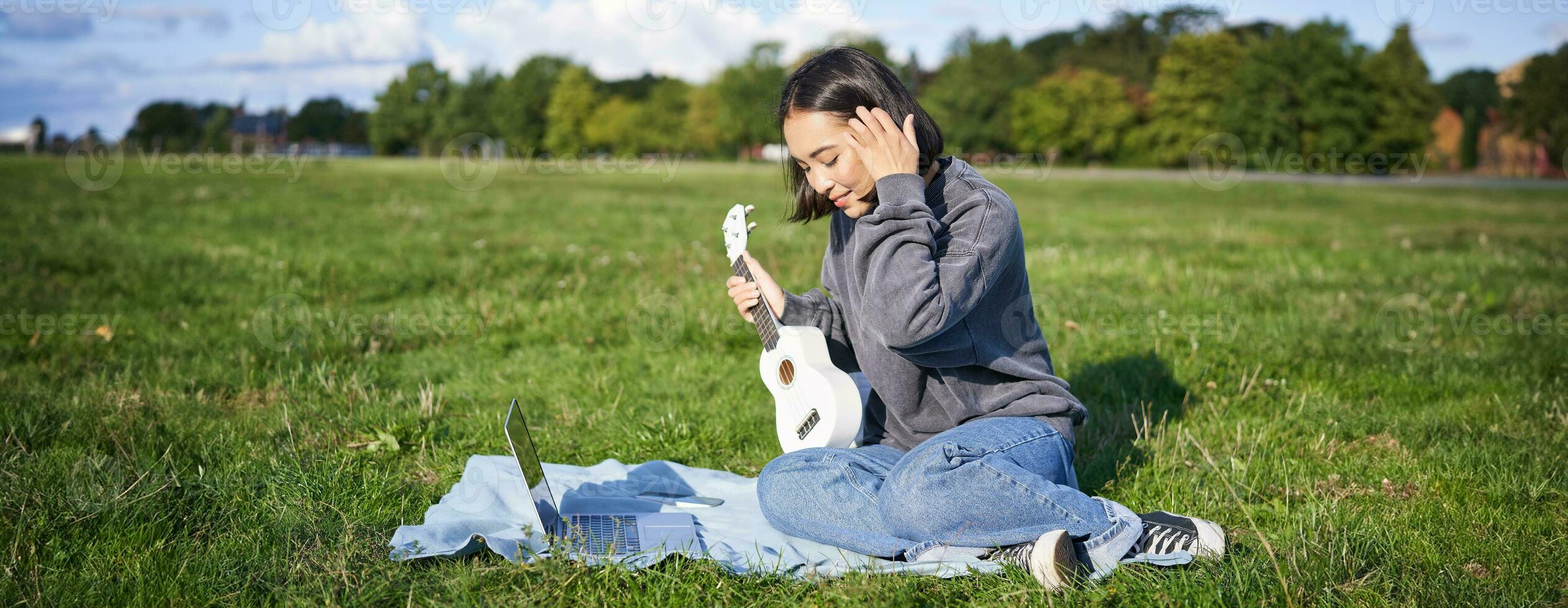 Smiling asian girl learns how to play ukulele via laptop, online video tutorials, sitting on grass in park with musical instrument photo