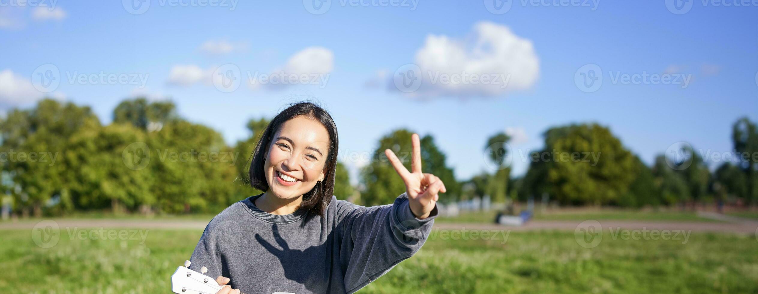 Vertical shot of positive asian girl shows peace sign, plays ukulele in park, rests and enjoys the day. Copy space photo