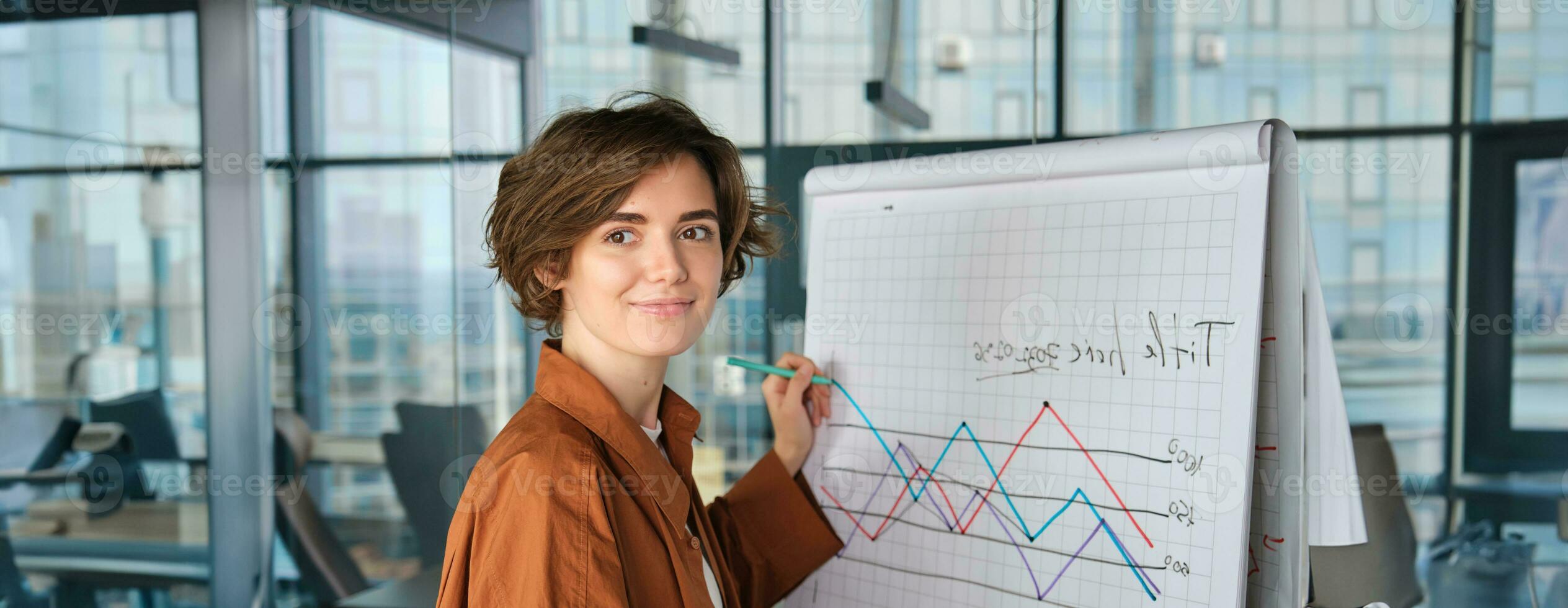 Image of young saleswoman, start up manager stands near board with diagram, giving presentation to team in office and smiling photo