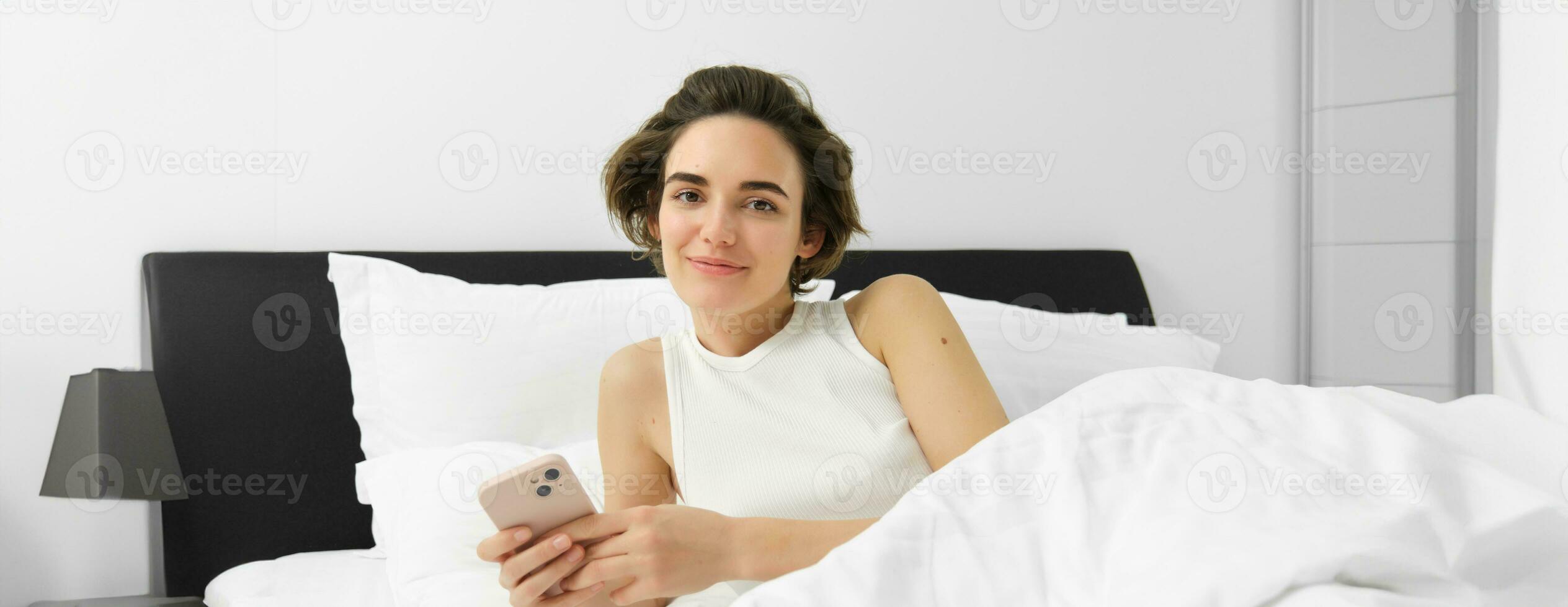 Portrait of young woman waking up in bed, using smartphone, turn off alarm on mobile phone, lying in her bedroom on white linen sheets photo