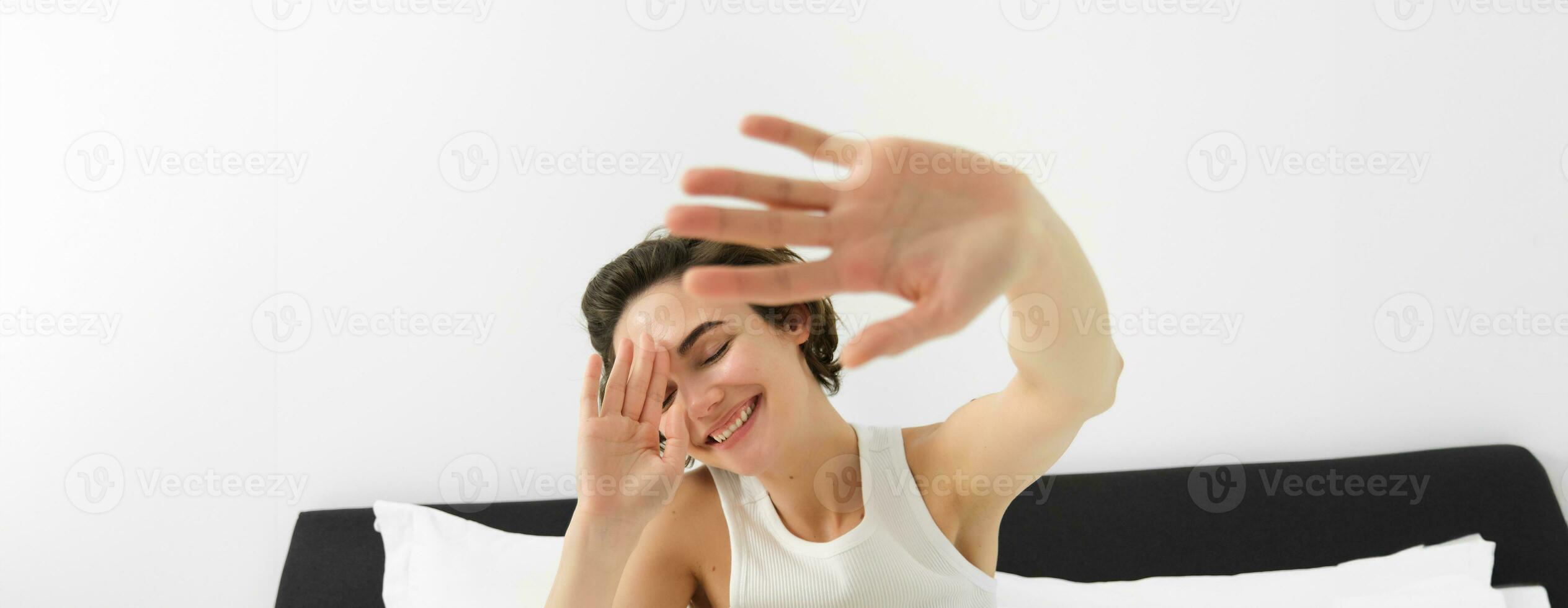 Portrait of sleepy, happy young woman, wakes up in her bed, stretching out hands, hiding face from camera, smiling and laughing photo