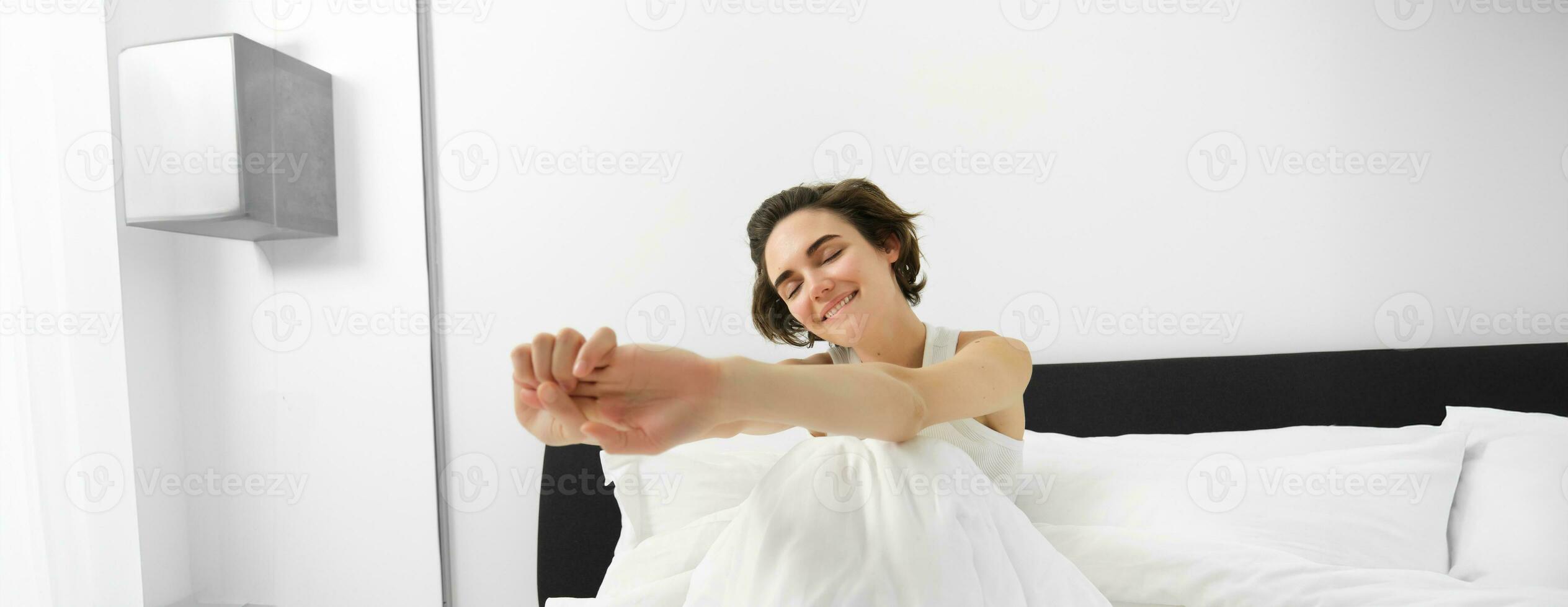 Portrait of smiling young woman wakes up in her bed, sitting under white linen sheets with closed eyes and stretching her hands with satisfaction photo