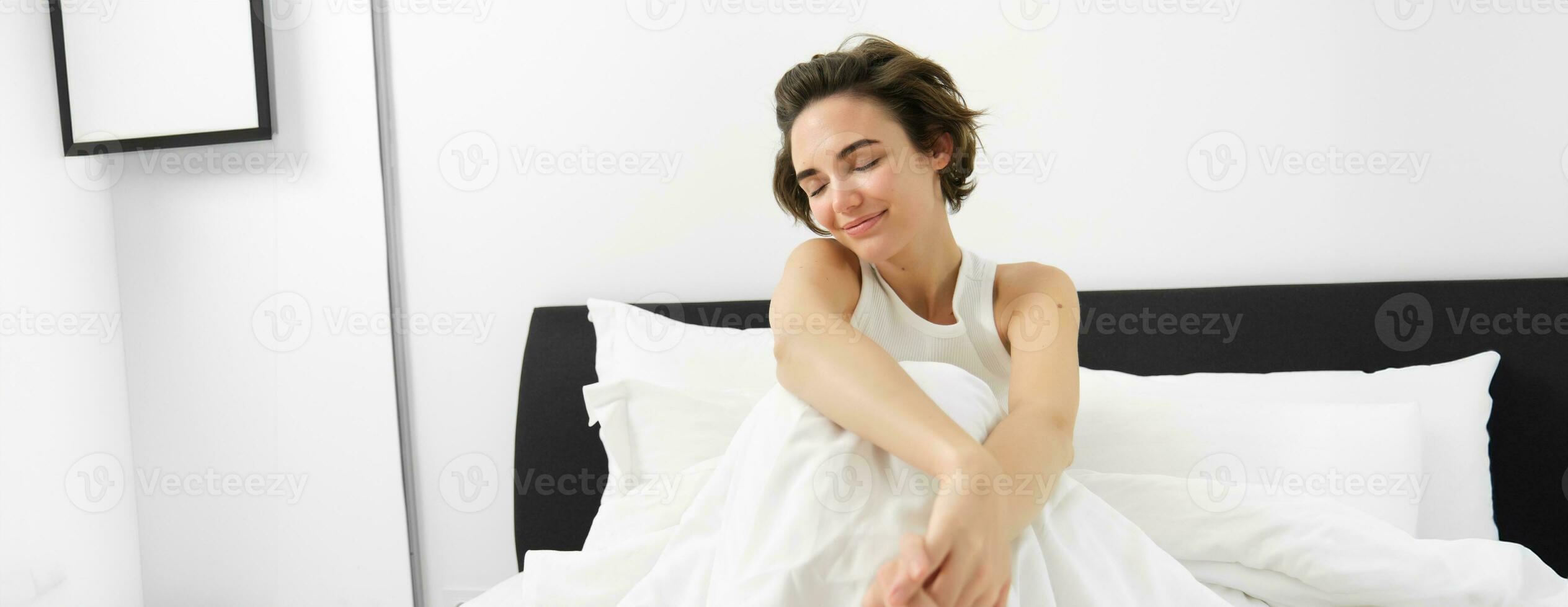 Portrait of cute smiling woman, sitting in bed, resting at home, lying under white linen sheets, waking up in morning with happy, pleased face expression photo