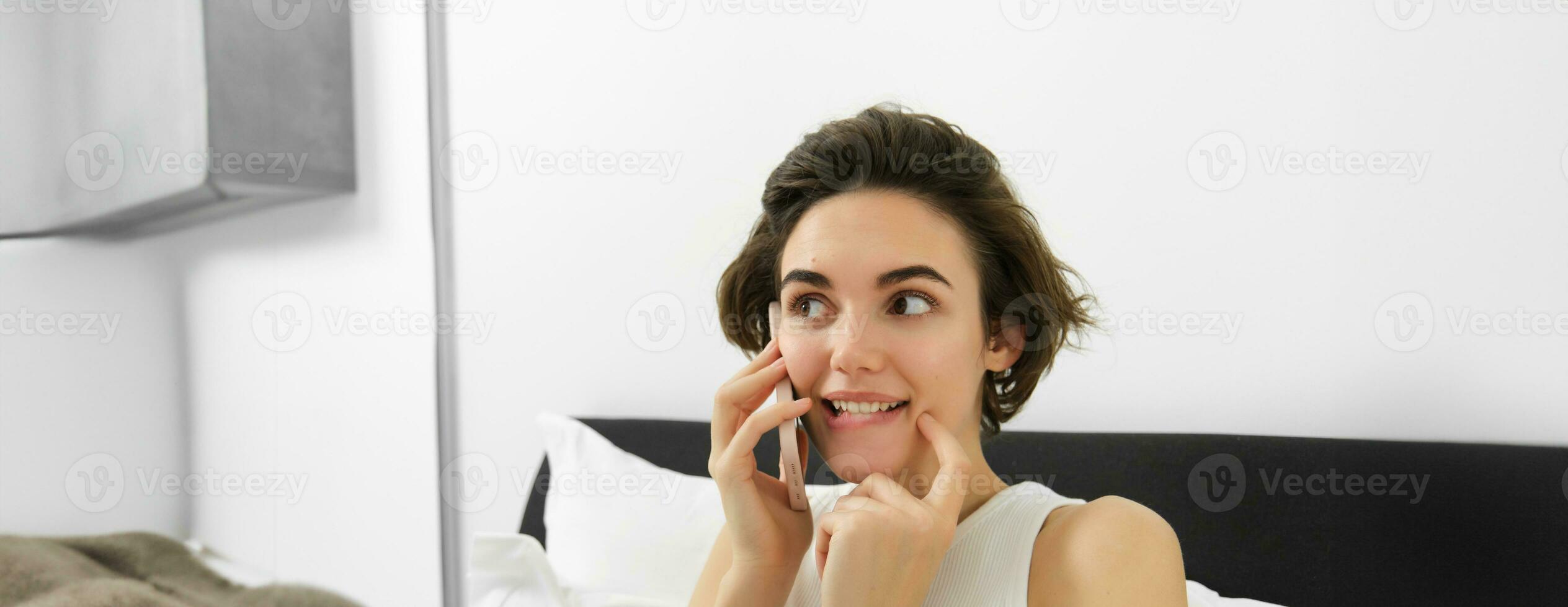 Close up of cute brunette woman, calling someone, looking excited, listening to friend over the phone, lying in bed photo