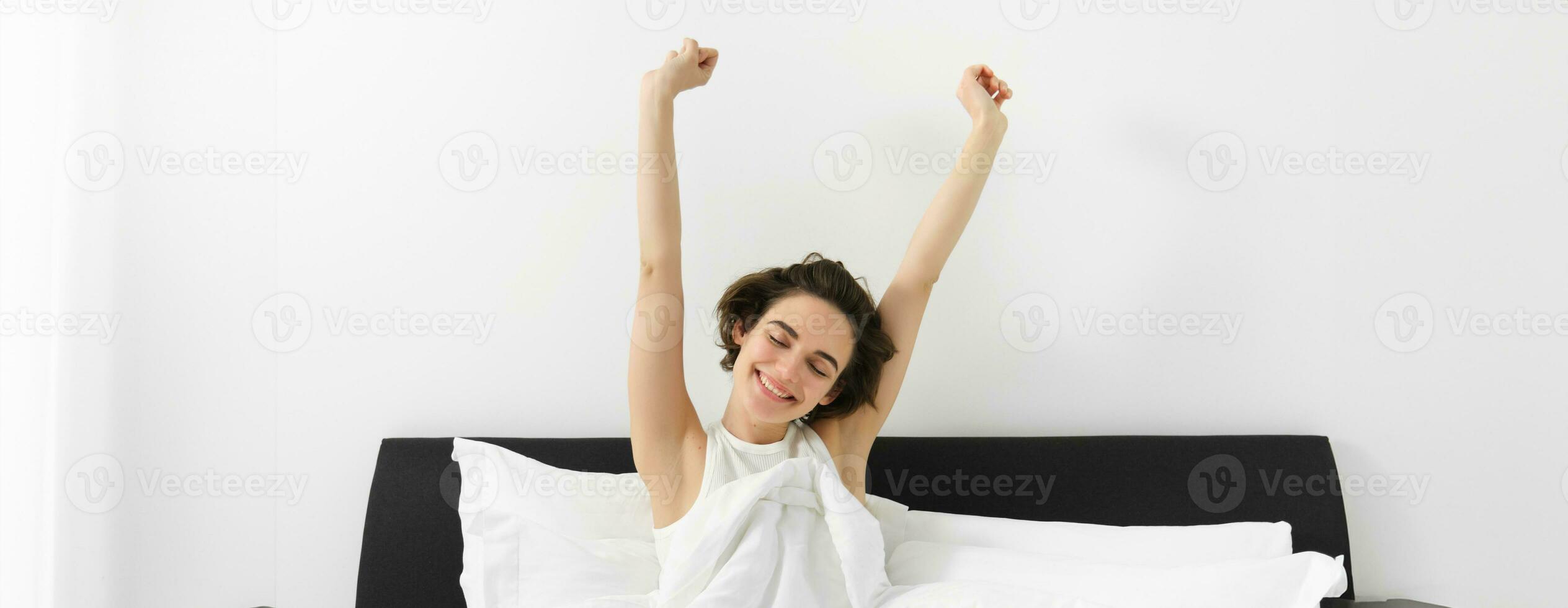 Beautiful young woman waking up in her bedroom, stretching hands with sleepy smiling face, sitting in bed under white linen duvet, had a good night sleep photo