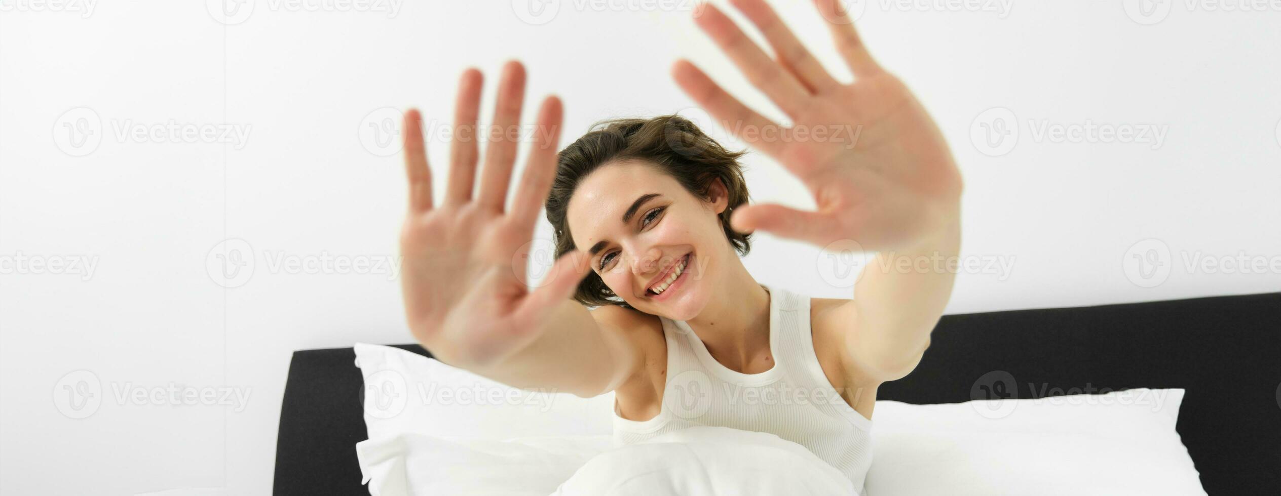 Portrait of sleepy, happy young woman, wakes up in her bed, stretching out hands, hiding face from camera, smiling and laughing photo