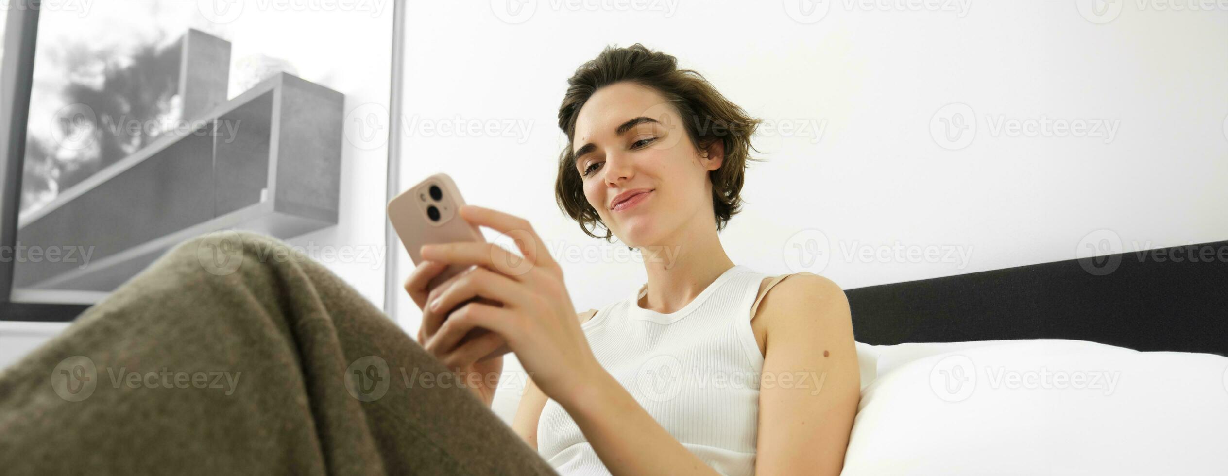 retrato de joven hermosa mujer acostado en cama, descansando en dormitorio, mensajería, utilizando móvil teléfono, participación teléfono inteligente y sonriente, desplazamiento social medios de comunicación, gasto comodidad hora a hogar foto