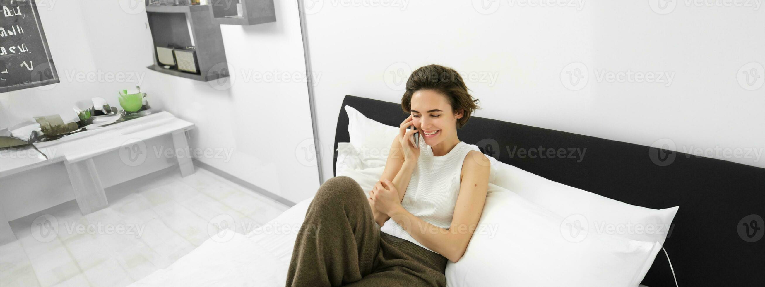 Beautiful young modern woman talking to friend, lying in bed and calling someone on phone, resting at home, chatting photo