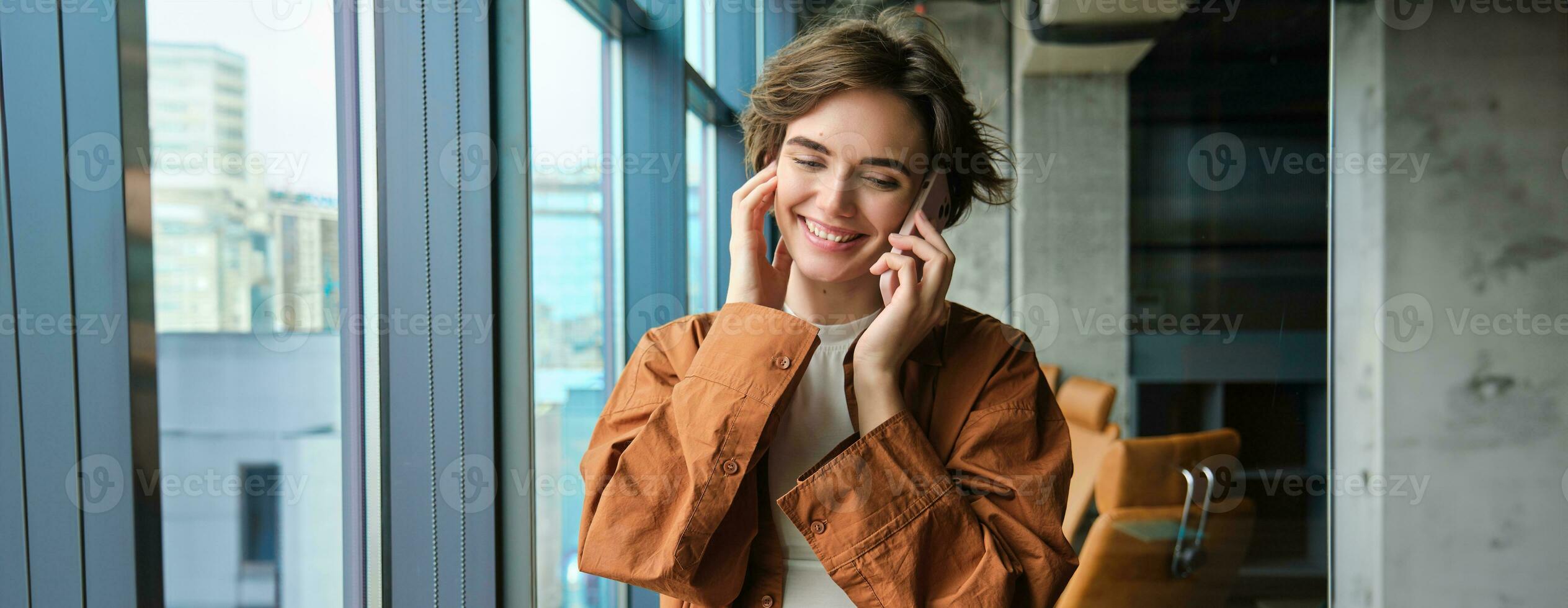 Portrait of working woman, social media manager in office, talking on mobile phone, having conversation on telephone, standing next to the window photo