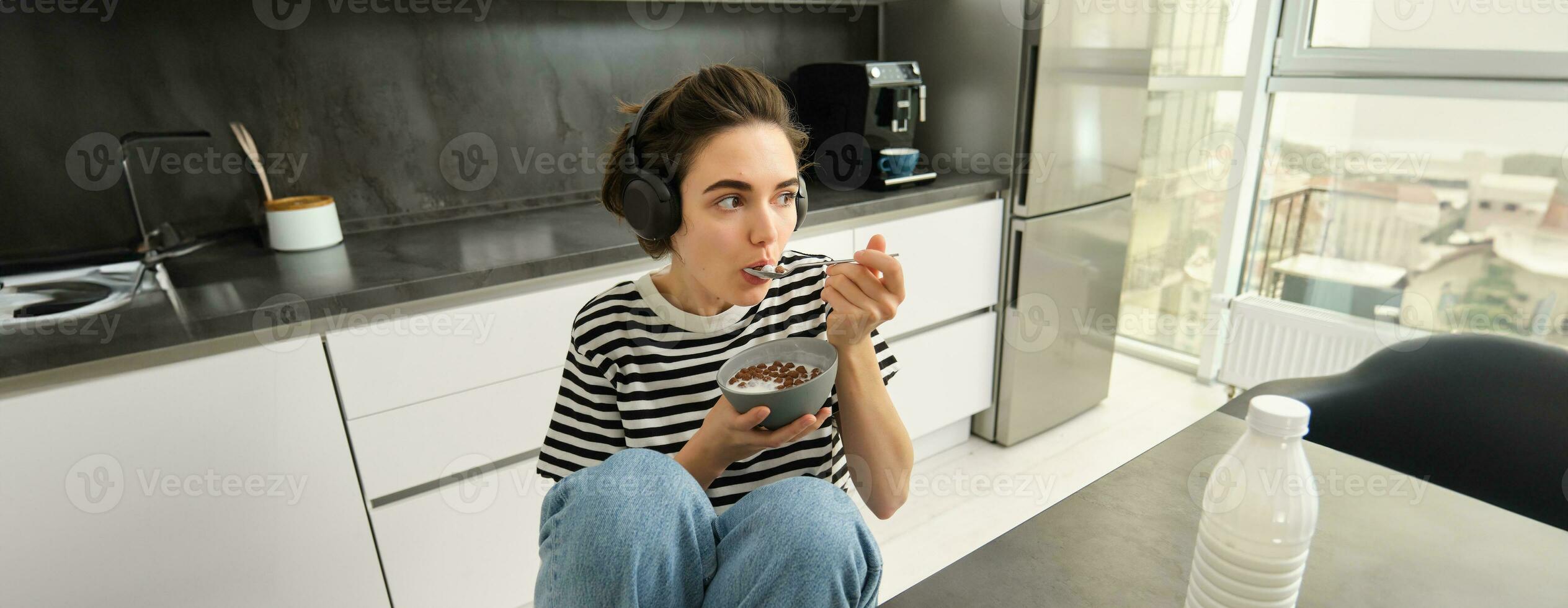 Image of stylish, modern woman, student eating cereals with milk for breakfast, holding bowl and spoon, listening music in wireless black headphones photo