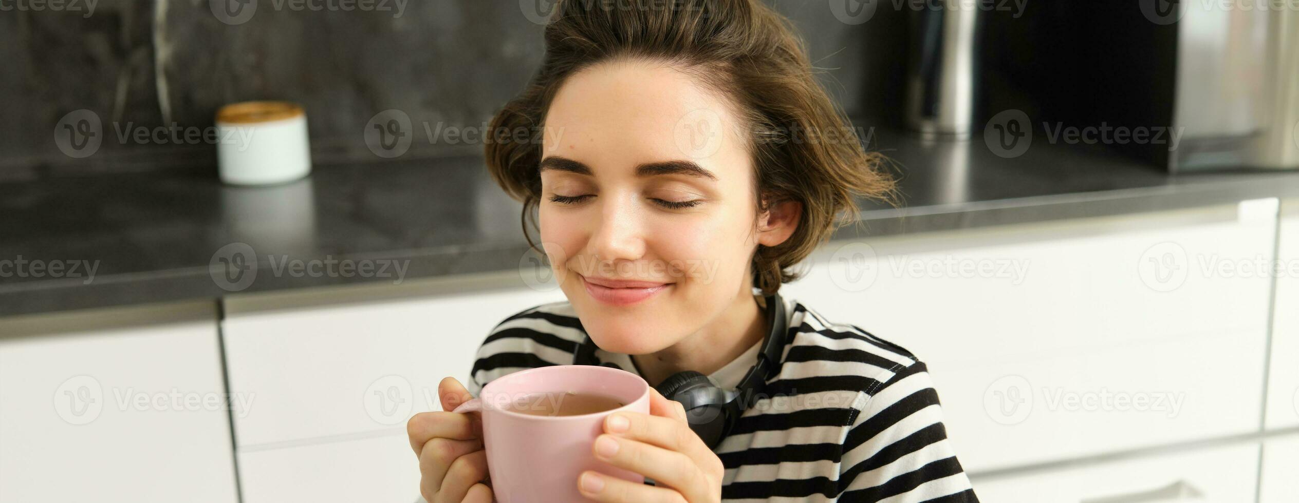 Close up portrait of woman with closed eyes and pleased smile, drinks warm tea with satisfaction, sitting in cosy kitchen photo