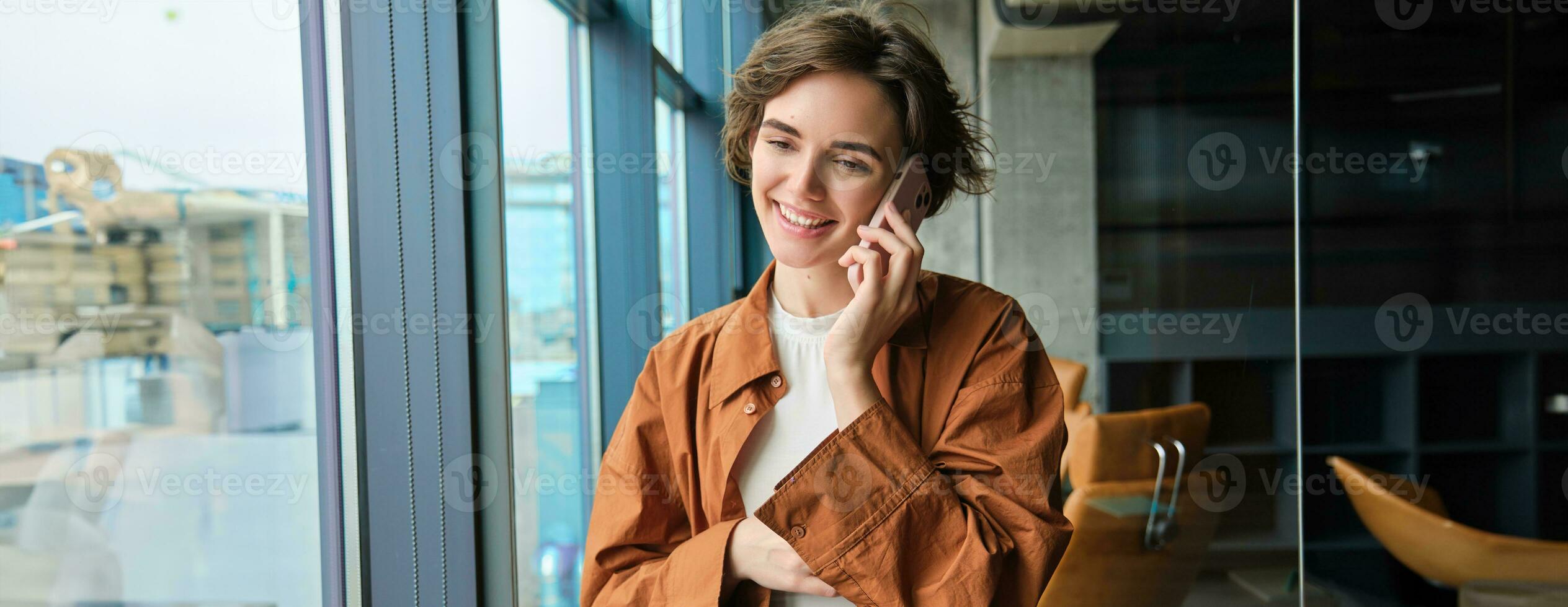 Happy young office worker near window, calling someone, answers a call on mobile phone, talking and smiling photo