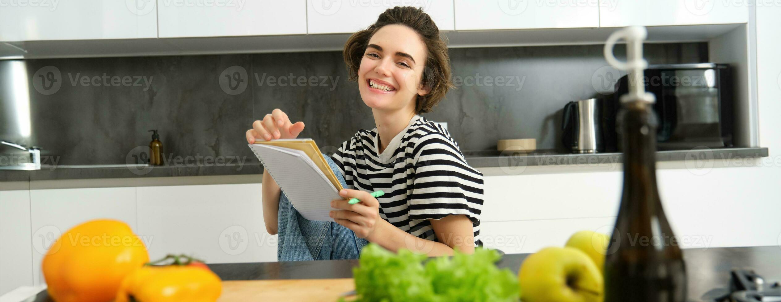 Portrait of young woman cooking, writing notes, grocery list in notebook, creating list of meals to cook through meal, sitting in kitchen near vegetables and chopping board photo