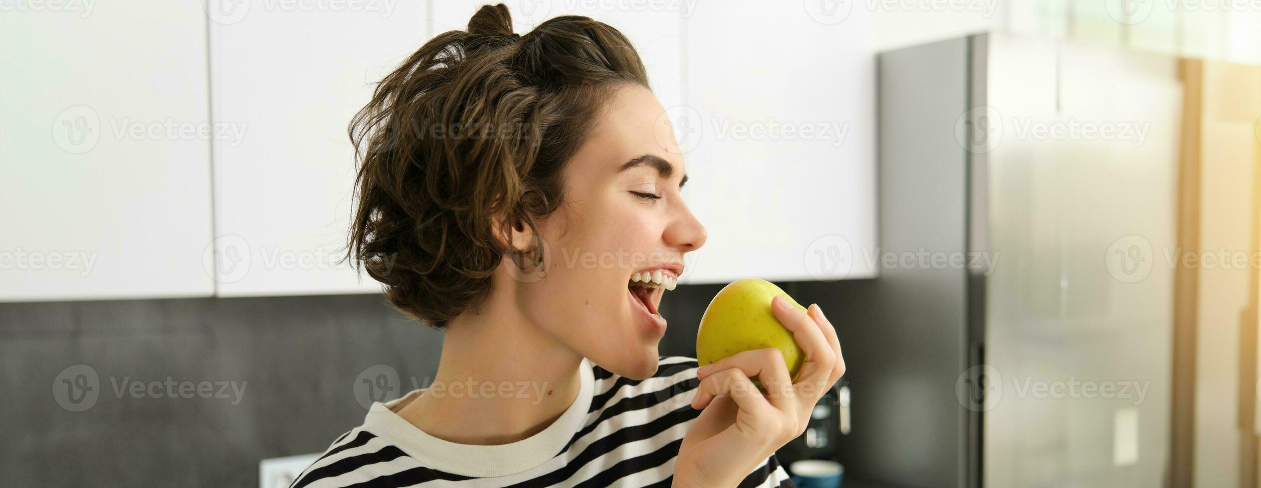 Close up portrait of young brunette woman biting an apple with pleasure, has pleased smile on her face, standing in the kitchen, having healthy snack for lunch, eating fruits photo