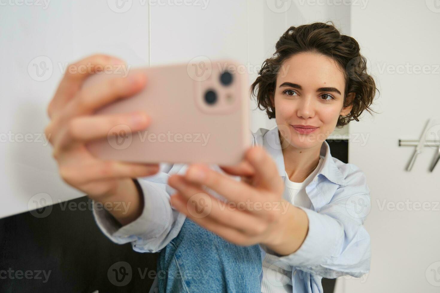 elegante joven niña bloguero, toma selfie en su cocina, poses para foto en móvil teléfono, utilizando cámara aplicación para un vídeo con filtros