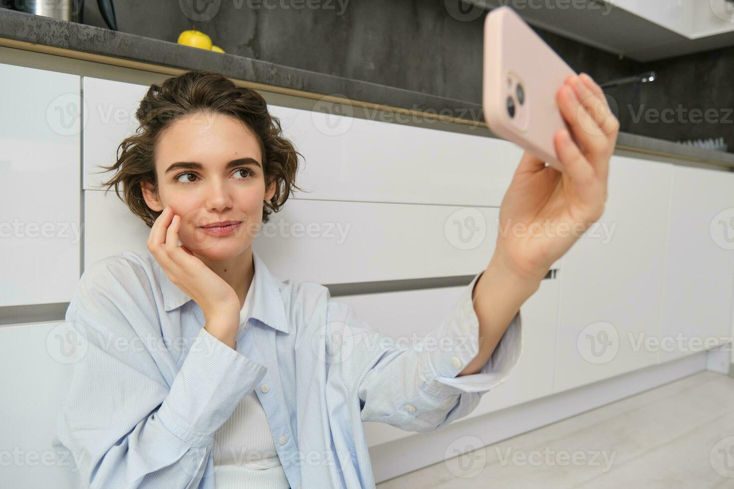 Portrait of young woman sits on kitchen floor with telephone, takes selfie on smartphone with app filters, poses for photo on mobile phone