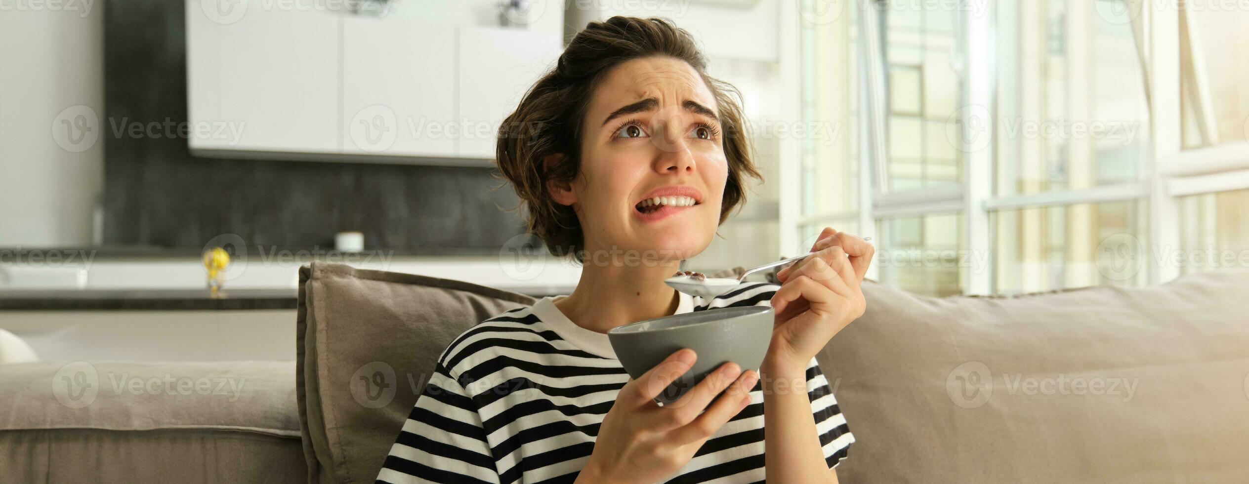 Close up portrait of female model, girl eating breakfast and watching intense scene in movie, staring at tv screen in living room, holding bowl of cereals with milk photo