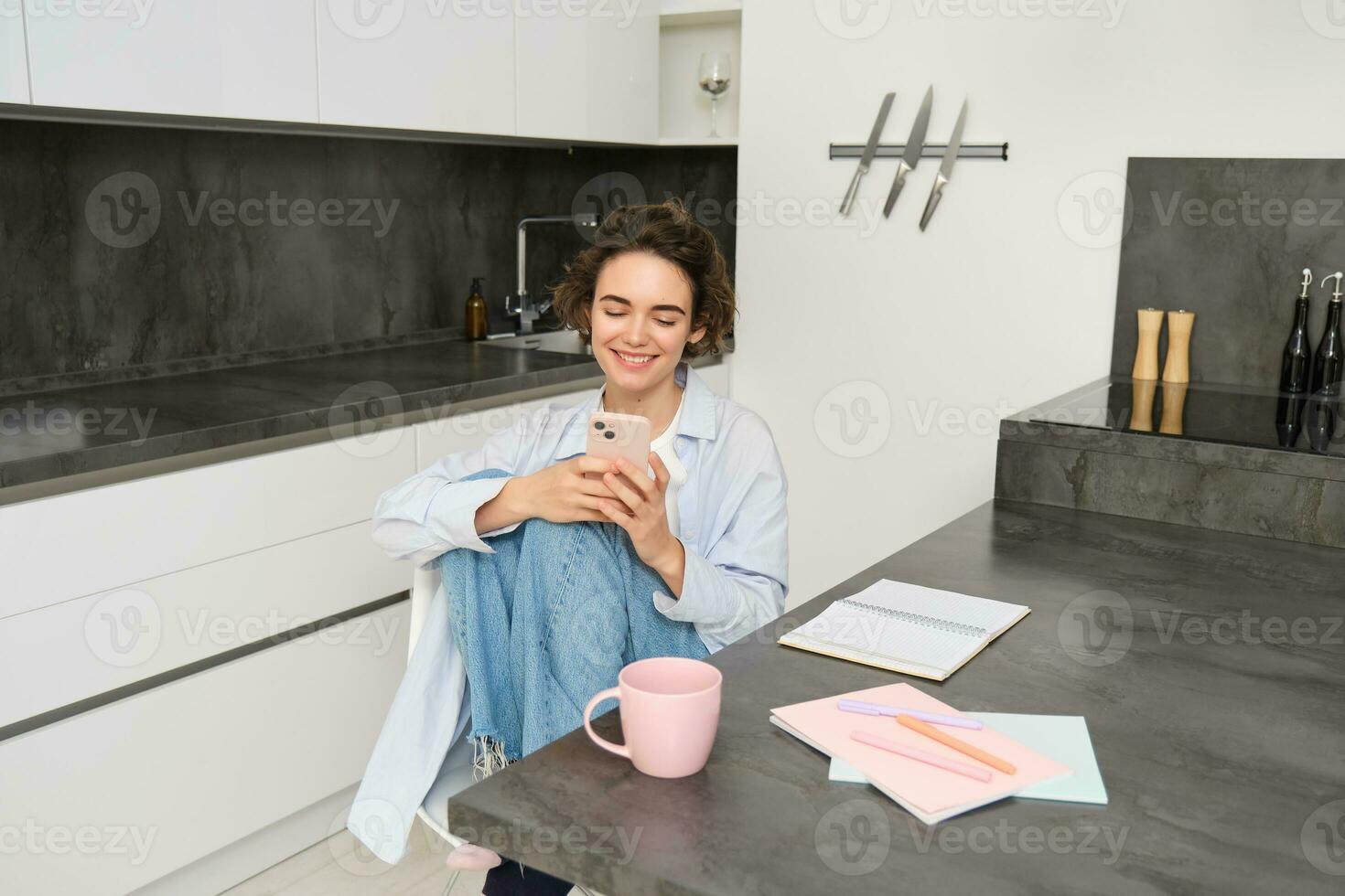 Young woman having break from work, sitting in kitchen and using smartphone, girl studies at home, sends a message on mobile phone photo