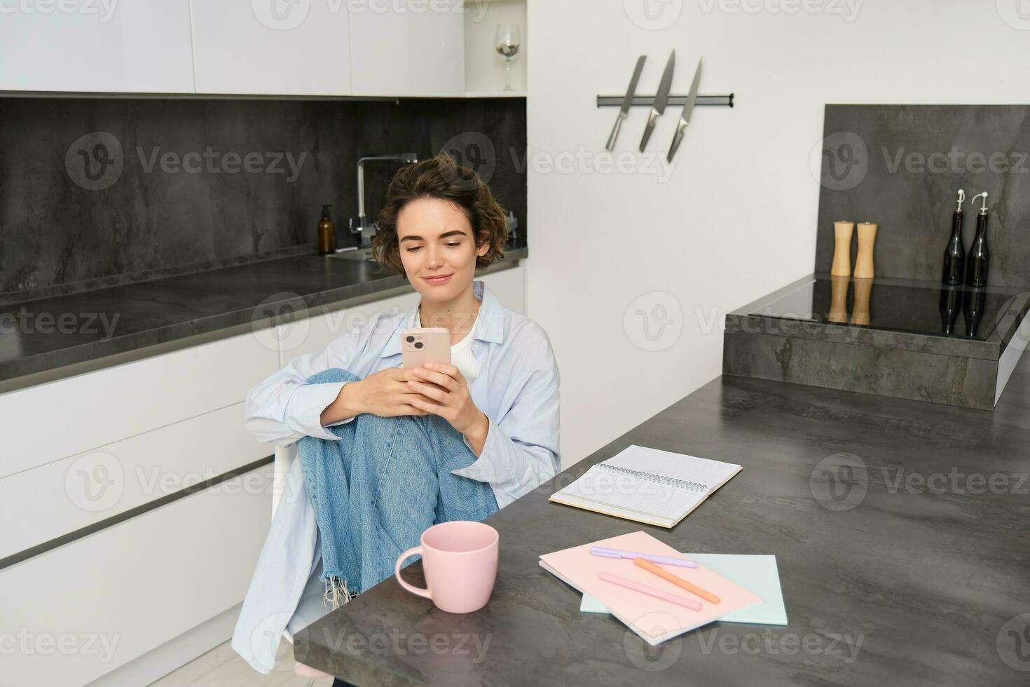 Young woman having break from work, sitting in kitchen and using smartphone, girl studies at home, sends a message on mobile phone photo