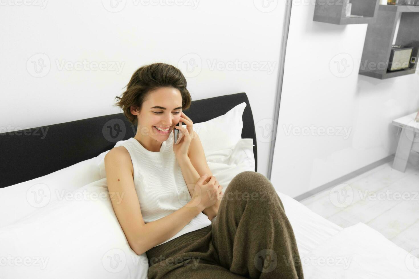 hermosa joven moderno mujer hablando a amigo, acostado en cama y vocación alguien en teléfono, descansando a hogar, chateando foto