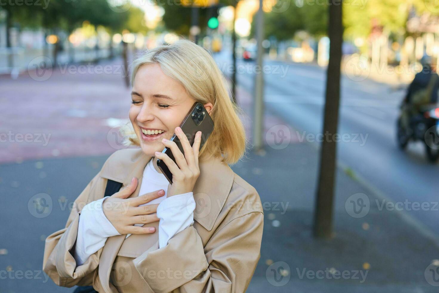 Close up portrait of beautiful young woman, blonge girl walking on street with mobile phone, chatting with friend, has happy face expression while talking over cellphone on her way photo