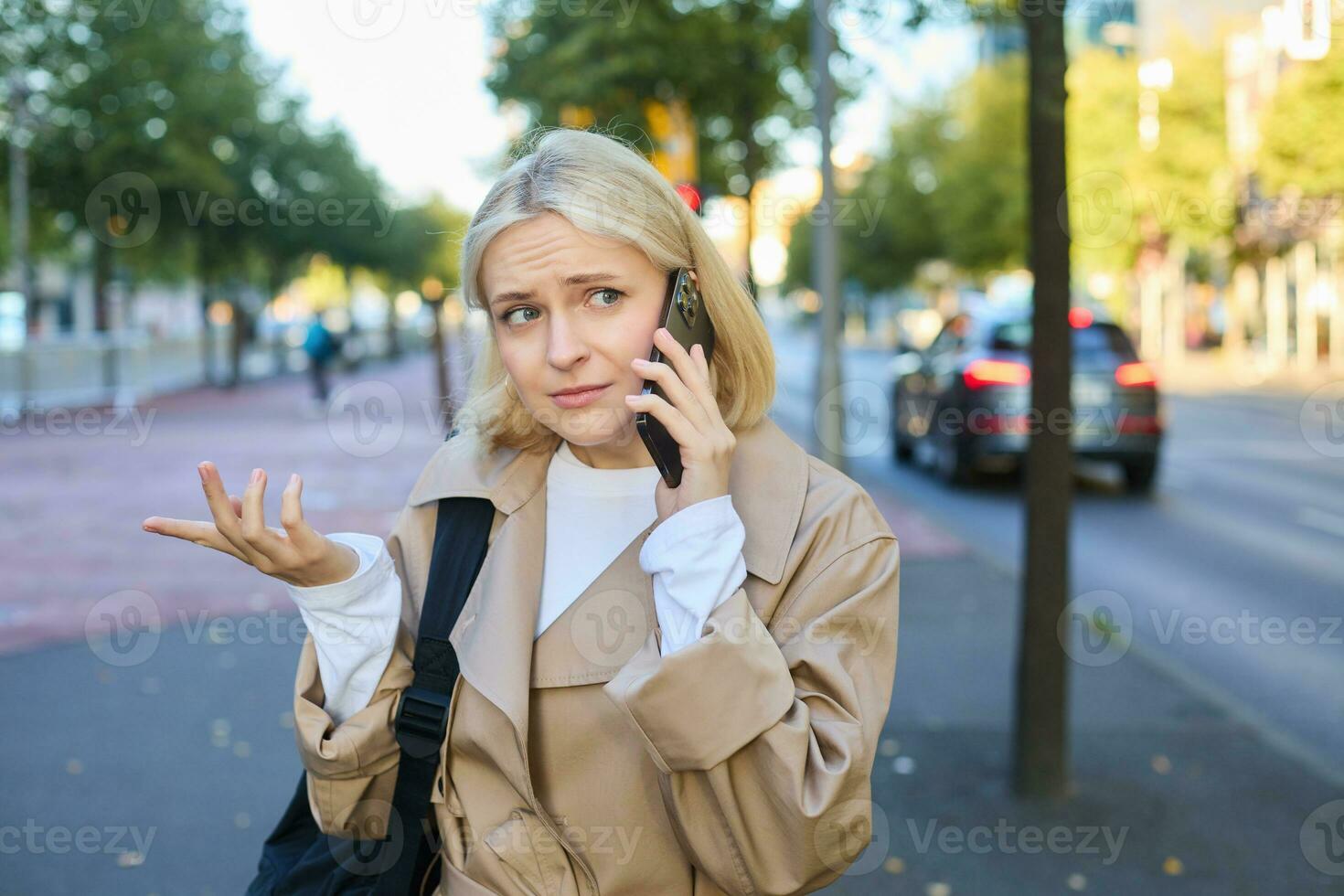 Portrait of woman with confused face, talking on mobile phone, answer phone call, standing on street with puzzled, clueless expression, posing on road photo