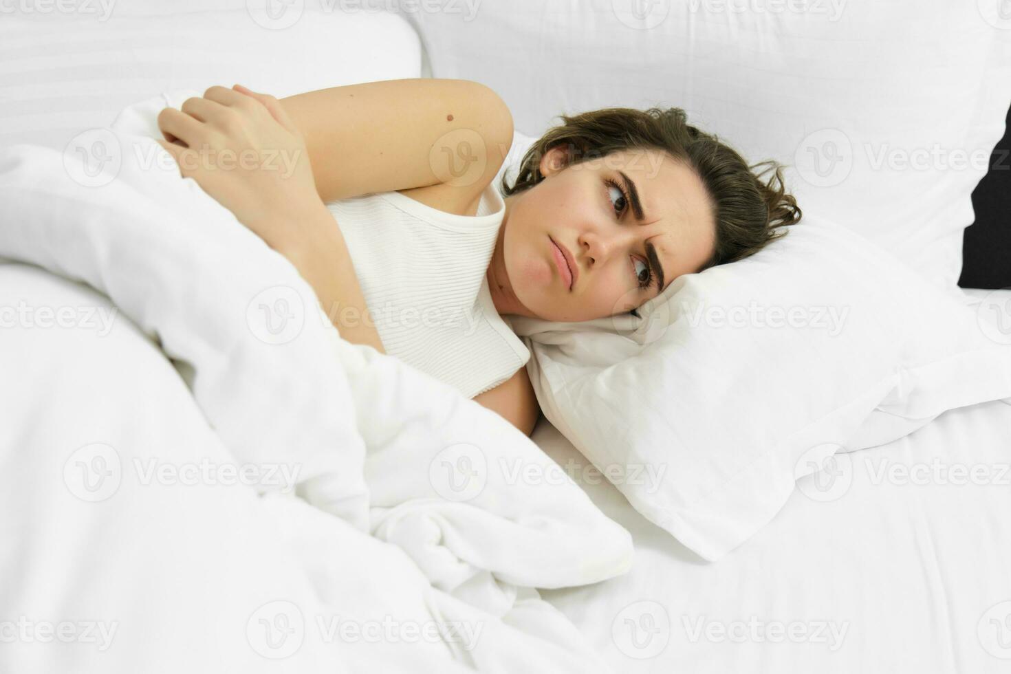 Close up of upset woman lying in bed with menstrual cramps, has period pain, touching her stomach and frowning, covered in white duvet photo