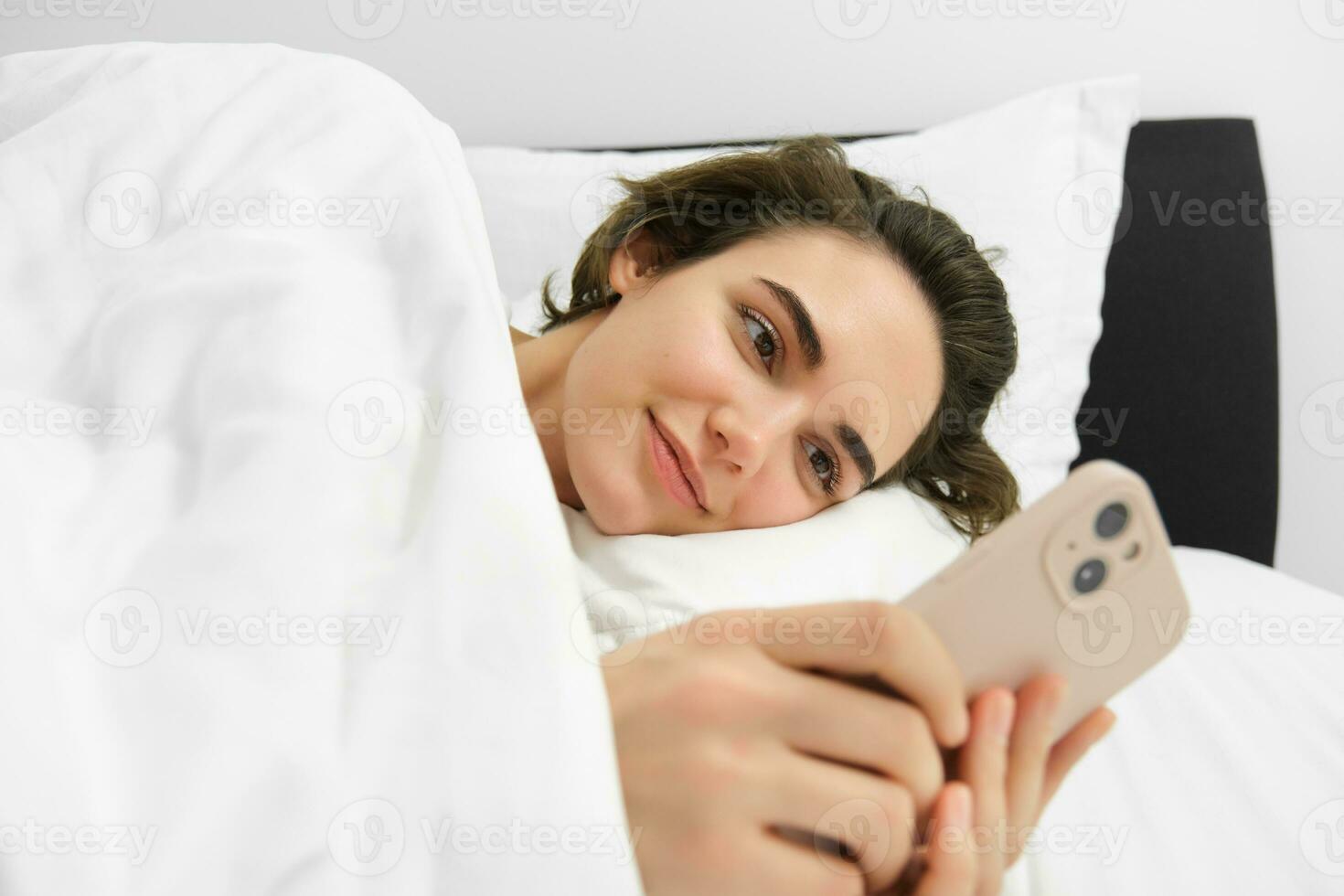 Smiling beautiful woman, lying in bed with smartphone, looking at mobile phone, using application, reading message and looking pleased photo