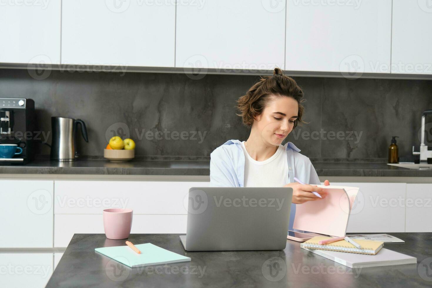 Portrait of working woman at home, checks her notebook, flips page, connects to online meeting via laptop. Girl learns programming remotely, studies course in internet photo