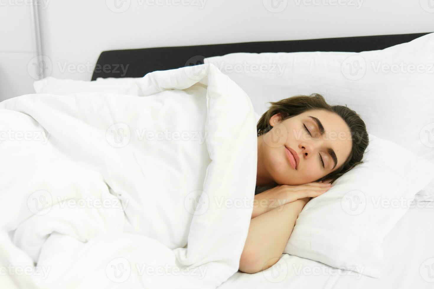 Image of beautiful young woman sleeping on comfortable bed, covered with soft white linen sheets, eyes closed, face is calm and unbothered photo