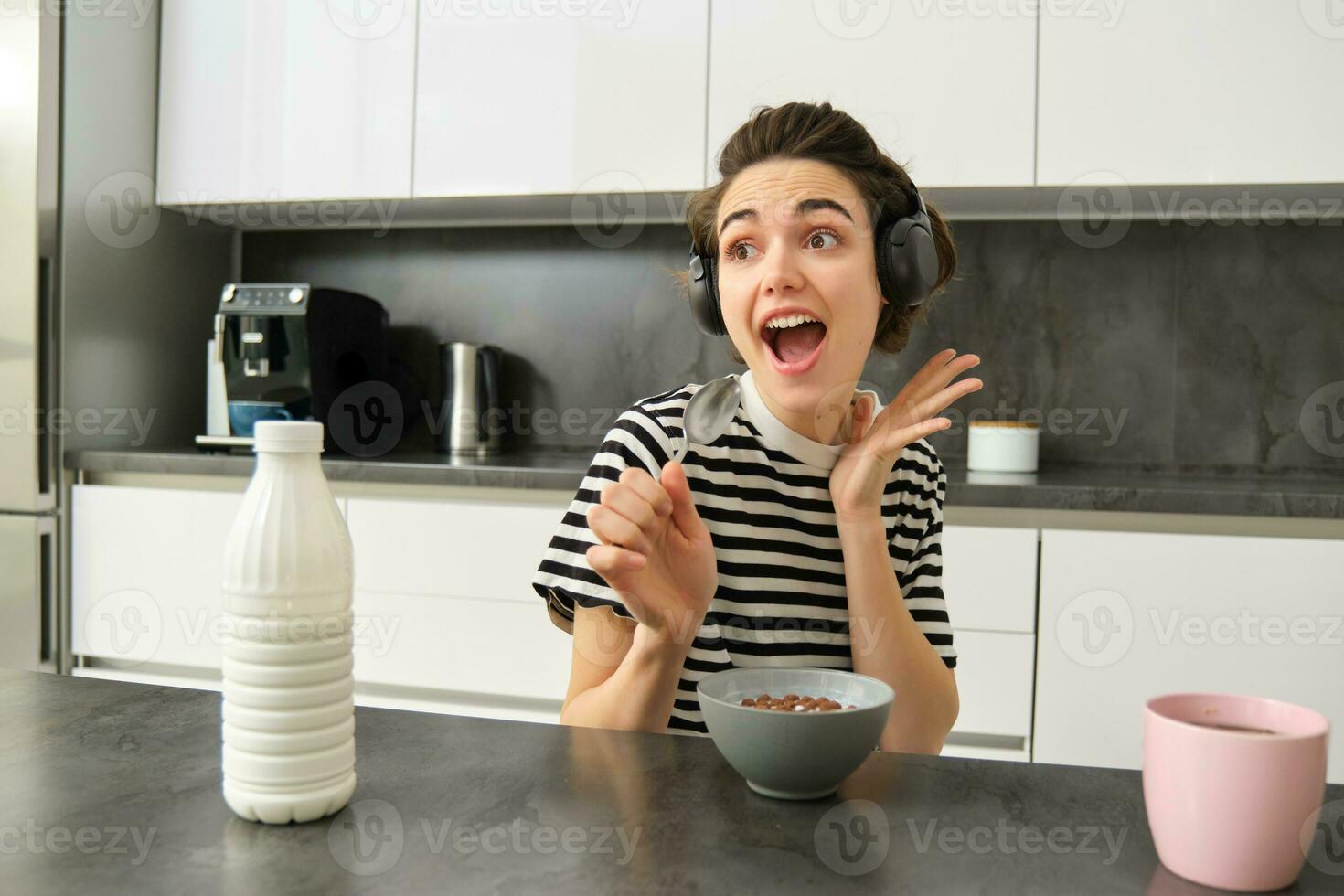 Happy and emotional young woman, singing while eating breakfast, having her cereals with milk, listening to music in wireless headphones, sitting in the kitchen photo