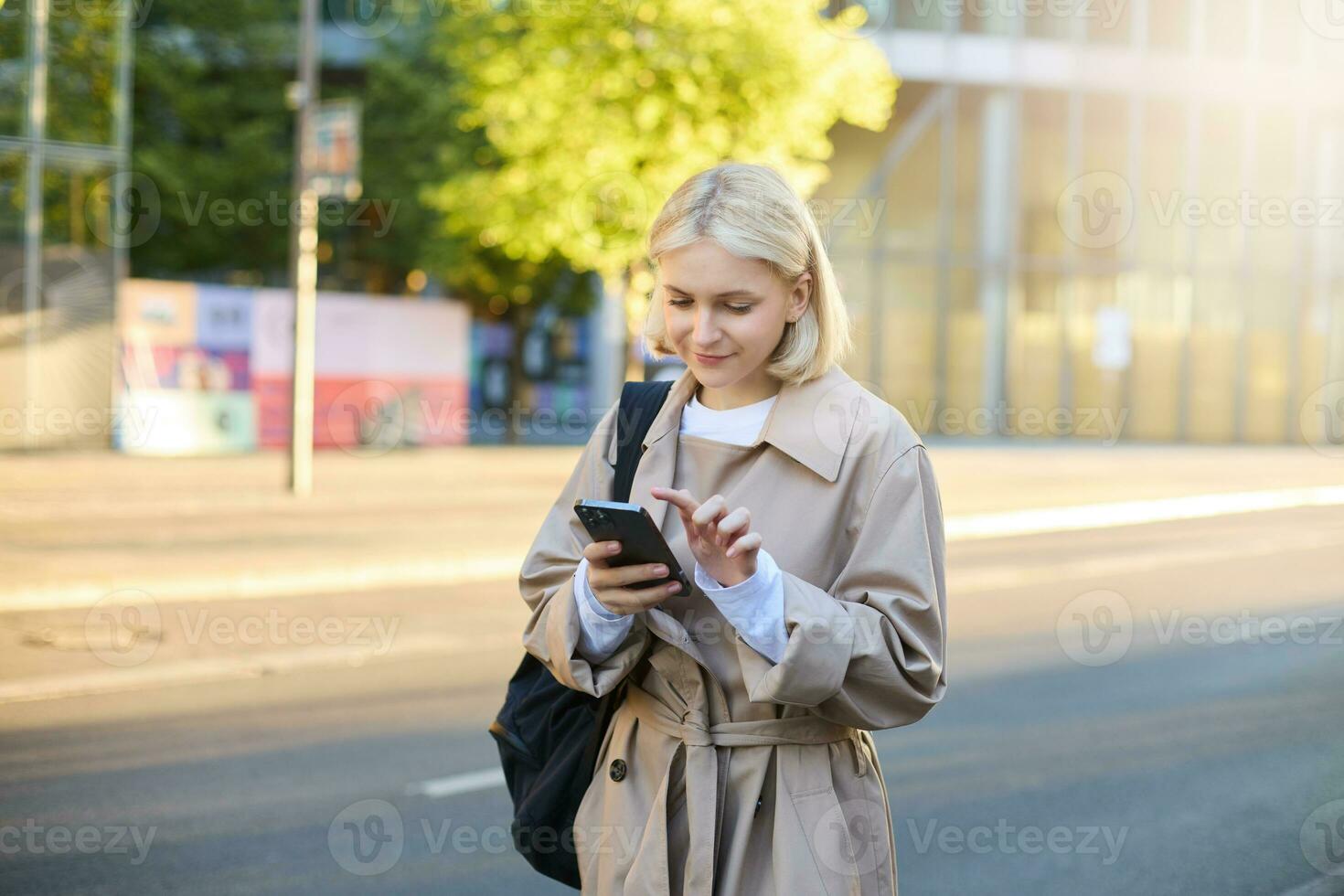 Image of young woman on street, carries backpack, looks at her smartphone, using carsharing application, orders taxi, sends a message on mobile app photo