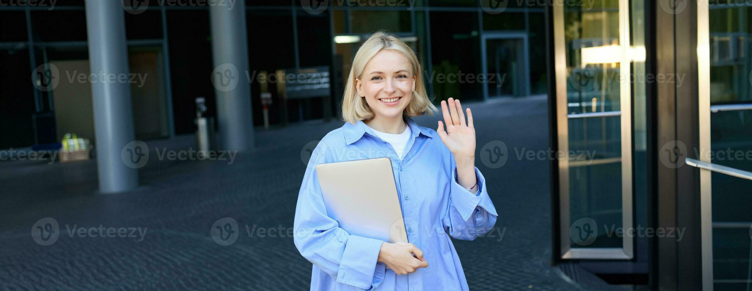 Friendly smiling student, blond girl with notebooks, tutor waving hand at camera, saying hello, standing near office building outdoors photo