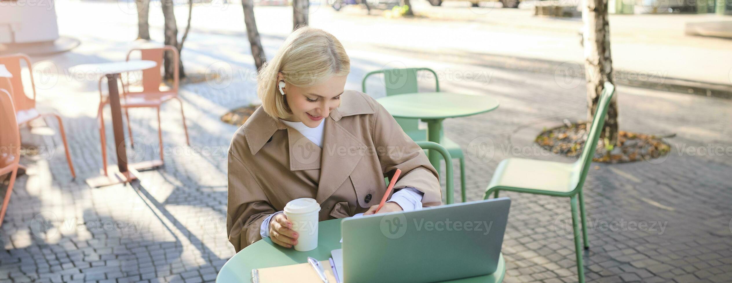 Portrait of young blond woman, female student in street cafe, wearing wireless headphones, using laptop, having online meeting, attend web lecture or course, doing homework photo