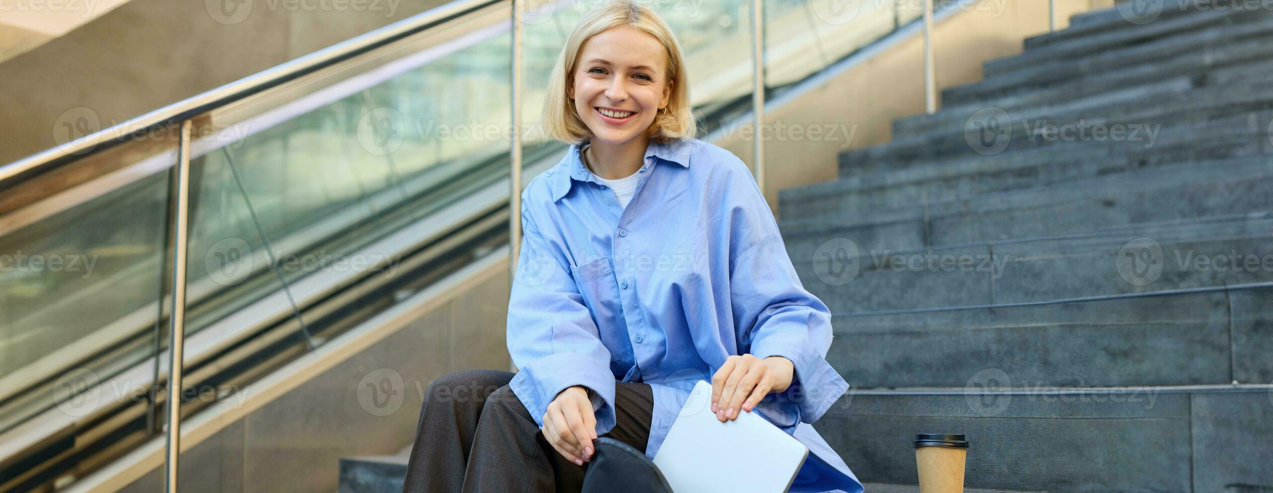 Portrait of young modern woman, student freelancer, sitting on public stairs outdoors, putting away her laptop, packing backpack, drinking coffee outside photo