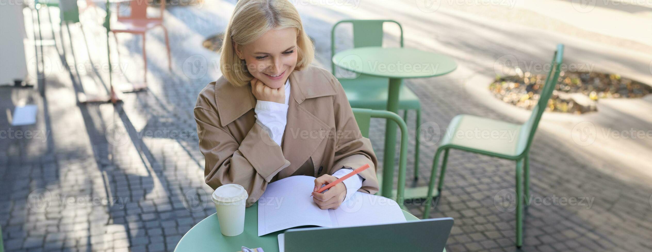 Image of young woman studying outdoors, sitting in cafe, making notes while listening to online lecture using laptop, making notes in notebook, writing photo