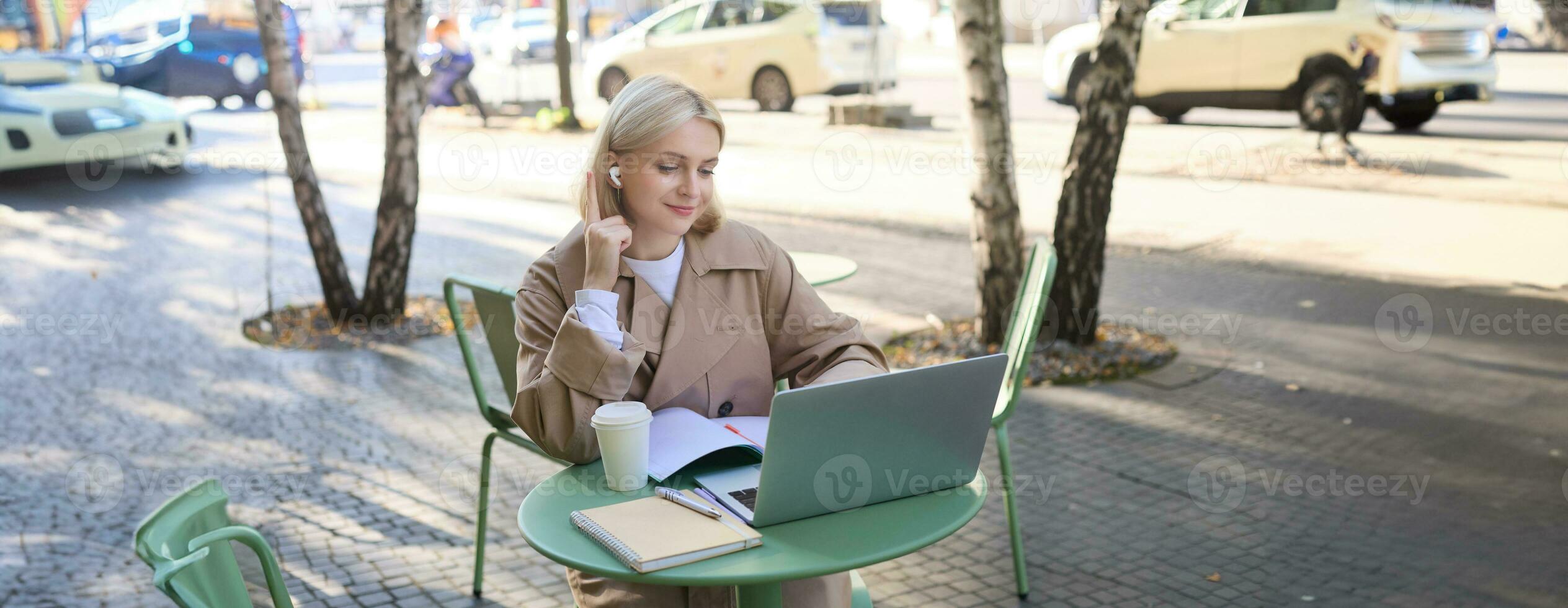 Young woman, freelancer, student doing homework in outdoor cafe, drinking her coffee on street, using laptop, connects to online lecture or doing course in internet photo