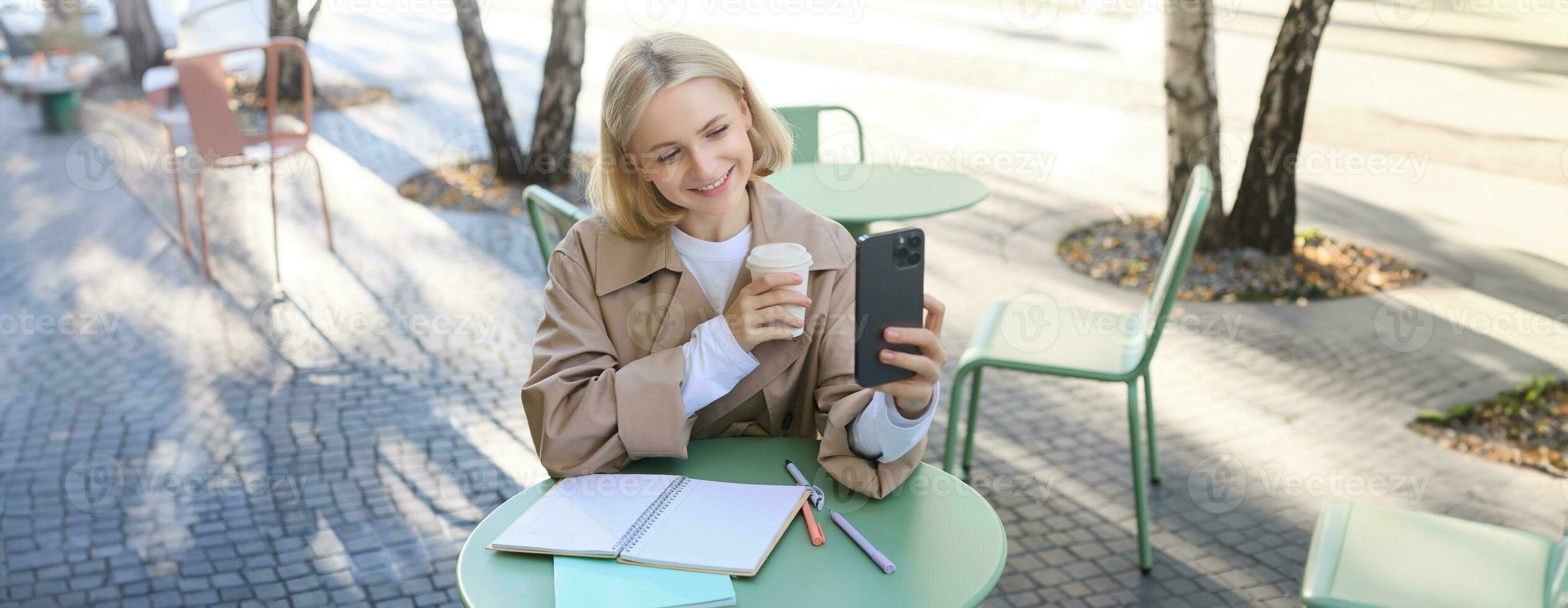 Image of stylish young woman, student taking selfie in cafe on street, posing with her favourite drink, enjoying coffee and making content for social media blog photo