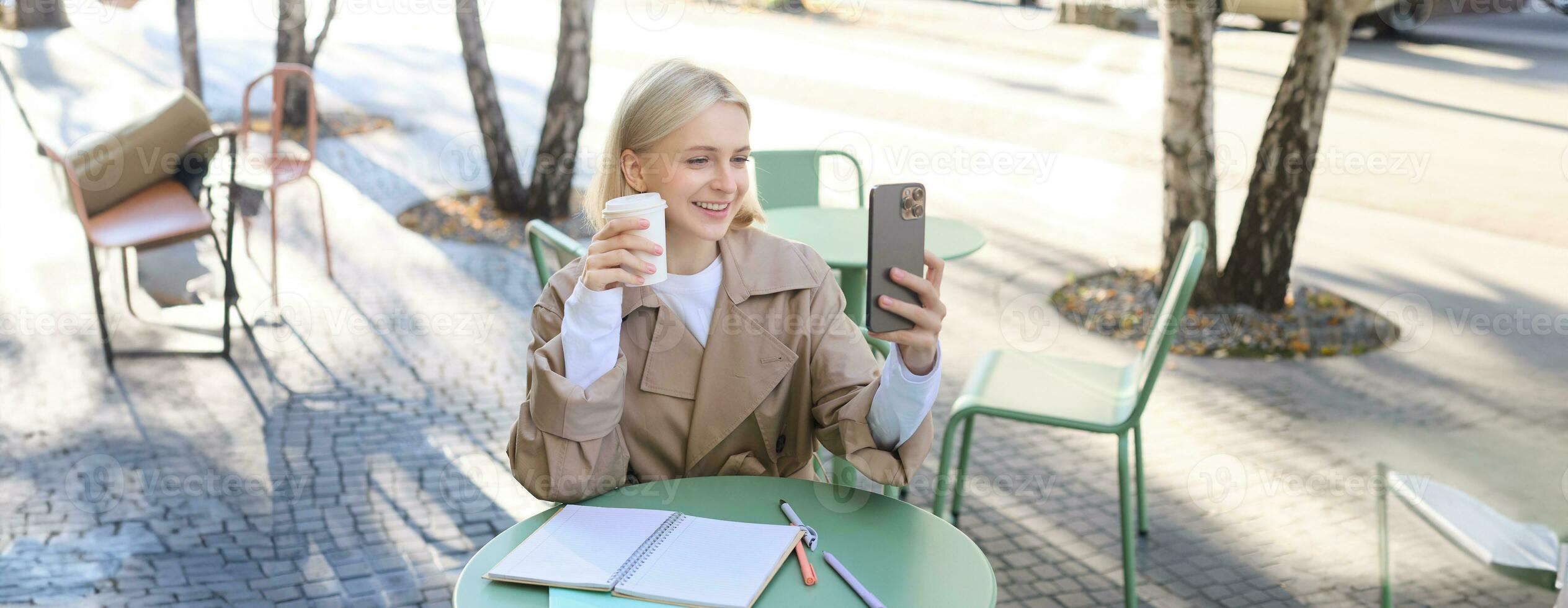 Image of stylish young woman, student taking selfie in cafe on street, posing with her favourite drink, enjoying coffee and making content for social media blog photo