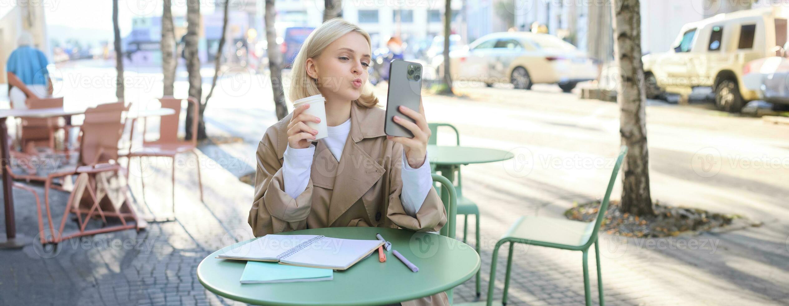 Image of stylish young woman, student taking selfie in cafe on street, posing with her favourite drink, enjoying coffee and making content for social media blog photo