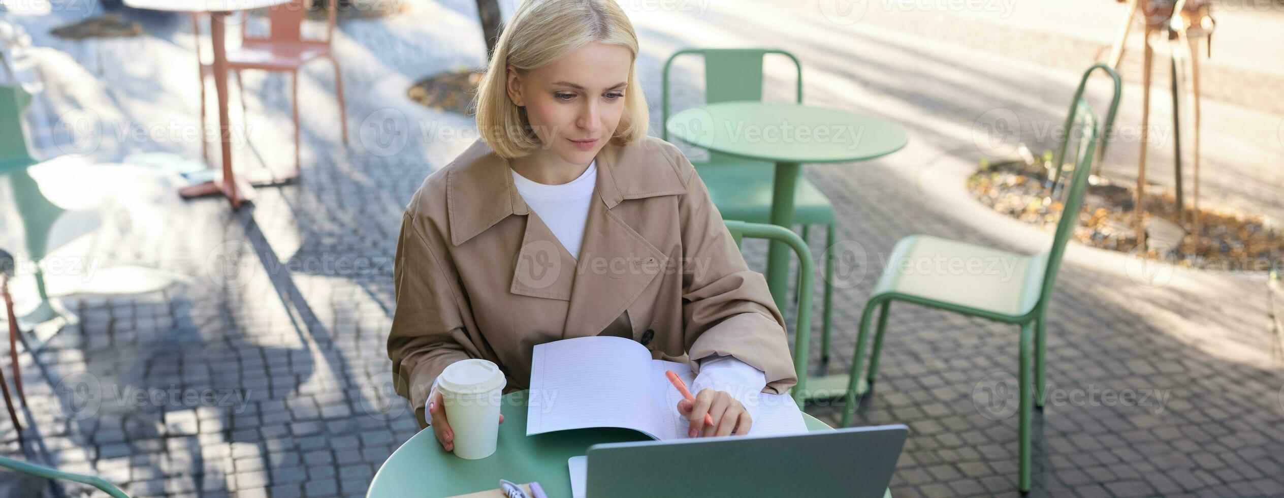 Image of young woman studying outdoors, sitting in cafe, making notes while listening to online lecture using laptop, making notes in notebook, writing photo