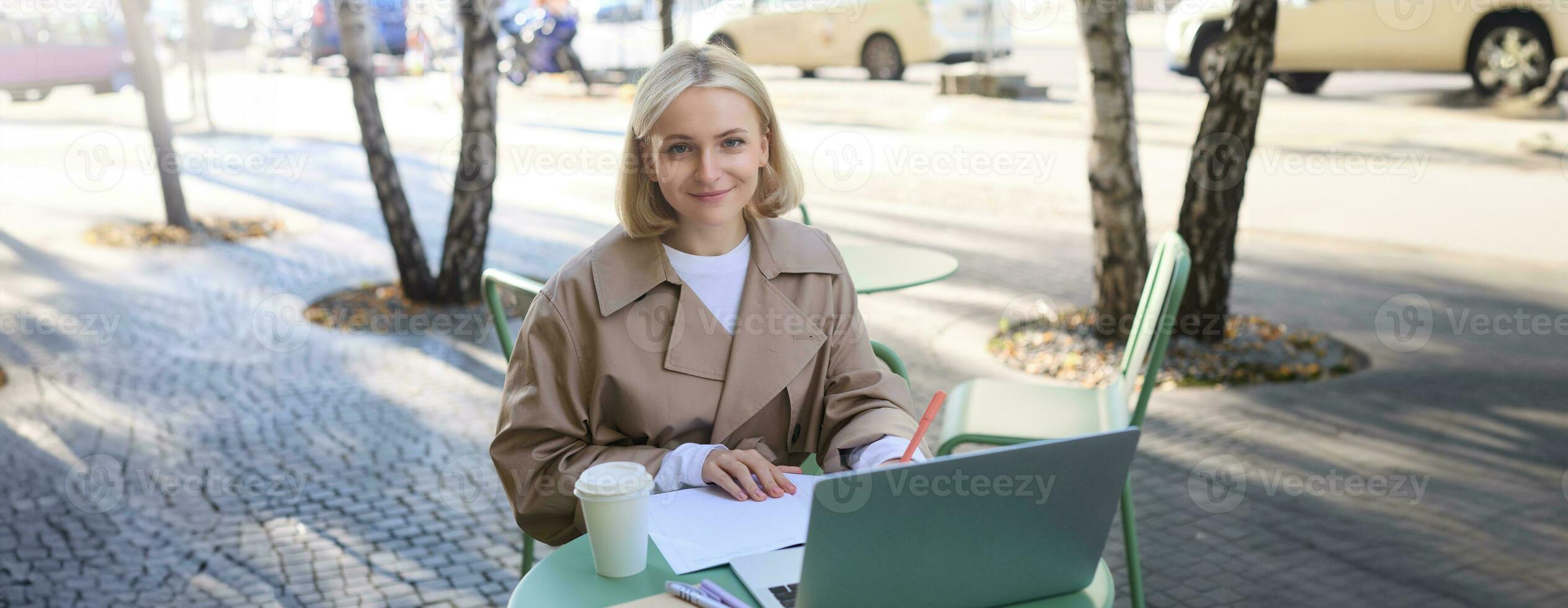 Image of young woman studying outdoors, sitting in cafe, making notes while listening to online lecture using laptop, making notes in notebook, writing photo