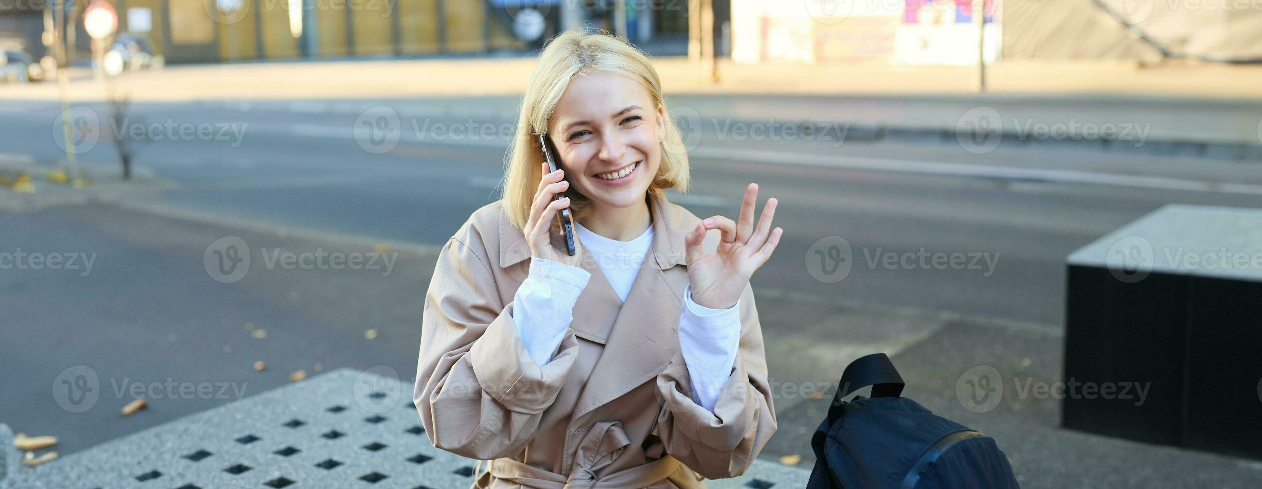 Happy beautiful young woman sitting on bench with mobile phone, talking on cellphone and showing okay gesture photo