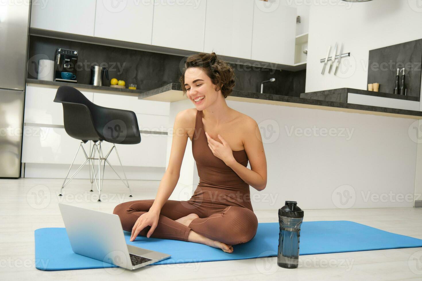 Sport and women. Young fitness instructor, yoga girl sits on rubber mat in her kitchen, waves hand and says hello at laptop, has online video gym class from home photo