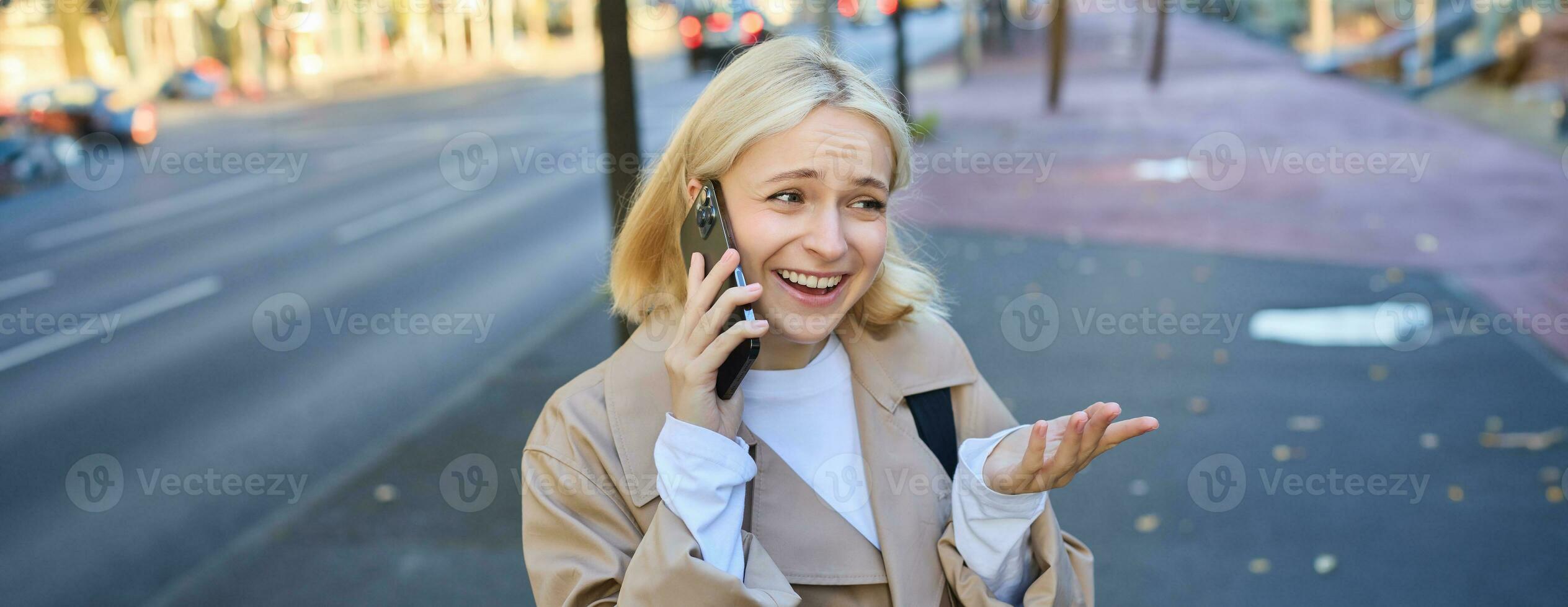 Lifestyle portrait of young blond woman talking on mobile phone, chatting with someone while walking on street, has conversation on smartphone and laughing photo