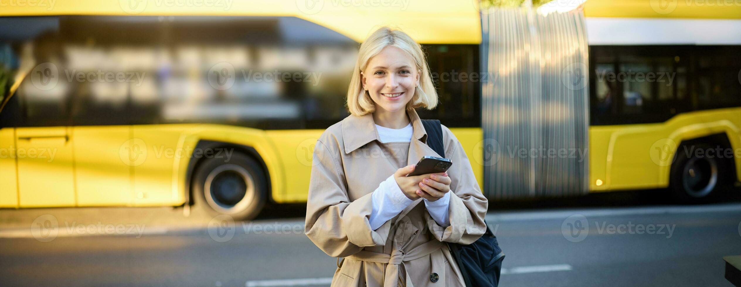 Outdoor shot of young woman with backpack, standing on street with cars passing behind her, holding mobile phone and smiling, urban lifestyle concept photo